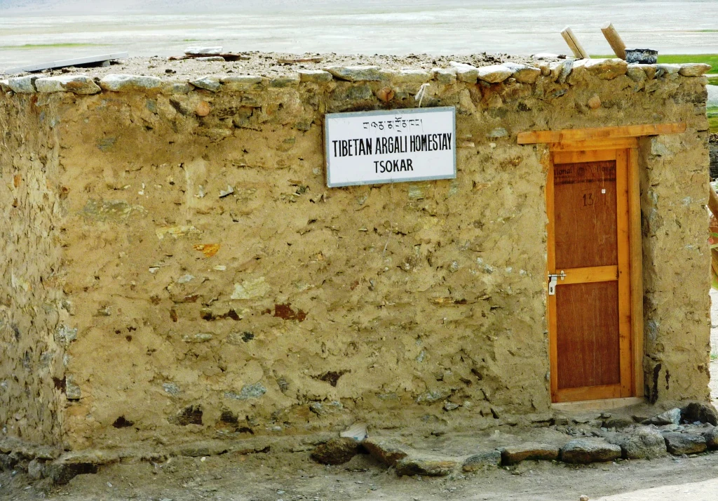 A small, single-story stone structure with a wooden door and a sign reading Tibetan Argali Homestay Tsokar mounted on the wall.