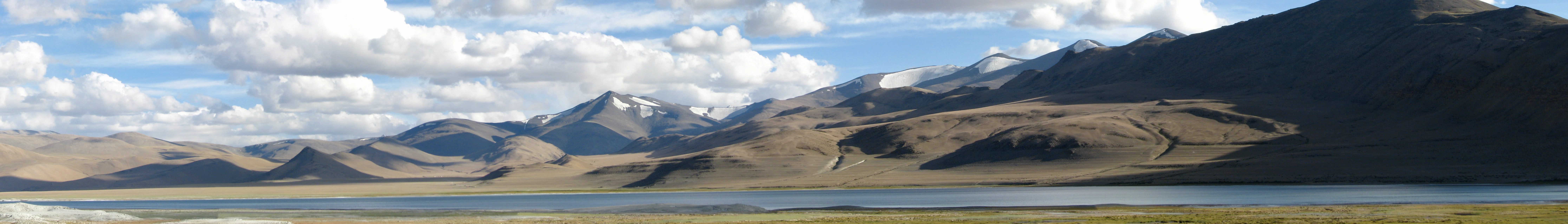 A wide panoramic view of a calm, blueish-grey lake in the foreground, with arid, brown, rolling mountain ranges extending across the background under a sky with scattered white clouds.