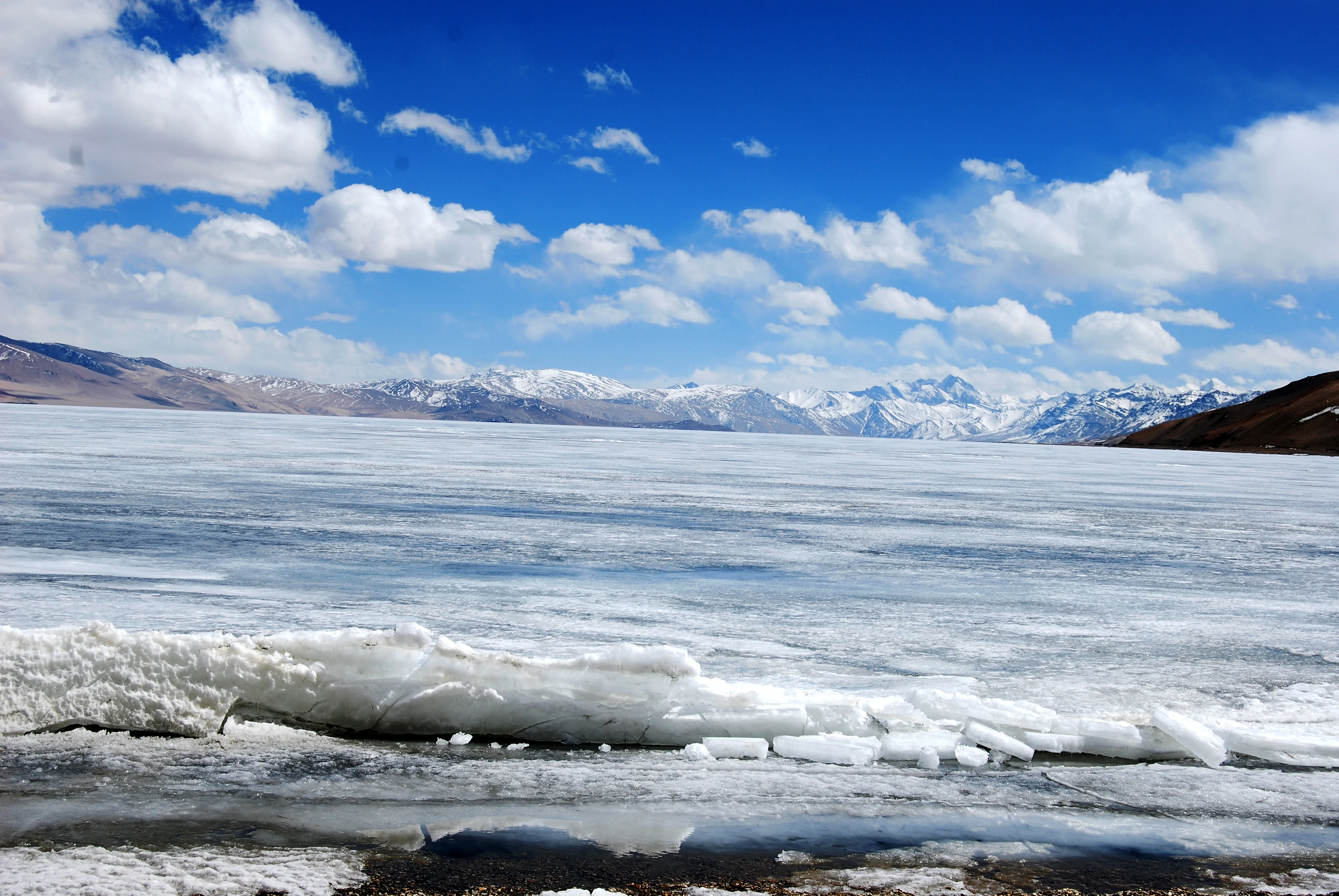 Una toma amplia a la altura de los ojos de un vasto lago de alta montaña congelado. El primer plano consiste en trozos de hielo roto a lo largo de la orilla, que conducen a una amplia extensión de hielo blanco congelado a través del lago. En la distancia, los picos de montañas nevadas se elevan bajo un cielo azul brillante lleno de nubes blancas dispersas.