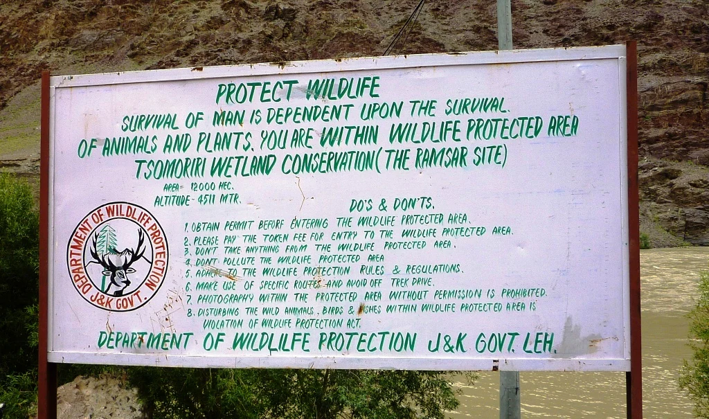 Un cartel metálico blanco con texto verde y el logotipo del Departamento de Protección de la Vida Silvestre del Gobierno de Jammu y Cachemira, que detalla las normas para la zona de conservación del humedal de Tsomoriri.