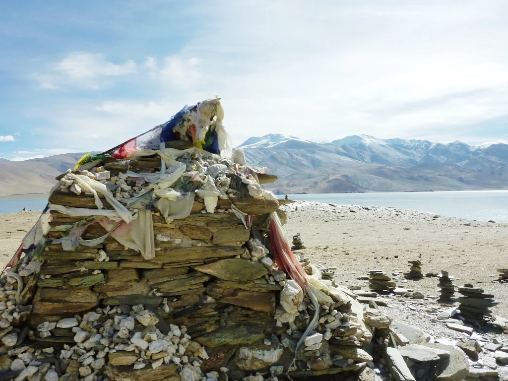 Un santuario budista de piedra coronado con coloridas banderas de oración situado en una orilla rocosa junto a un gran lago de alta altitud con montañas cubiertas de nieve en la distancia.