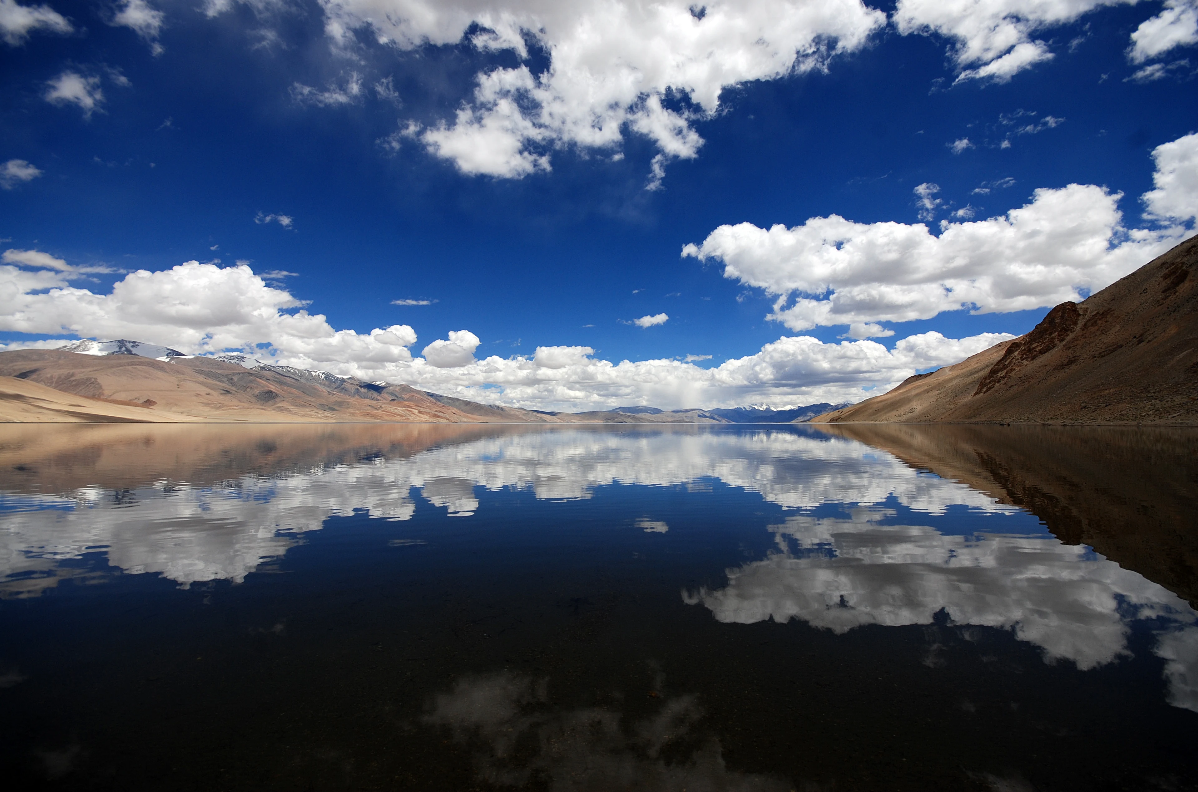 Una toma amplia y a baja altura del lago Kyagar con un claro reflejo de nubes blancas y cielo azul profundo en la superficie del agua, flanqueado por colinas montañosas marrones y áridas.