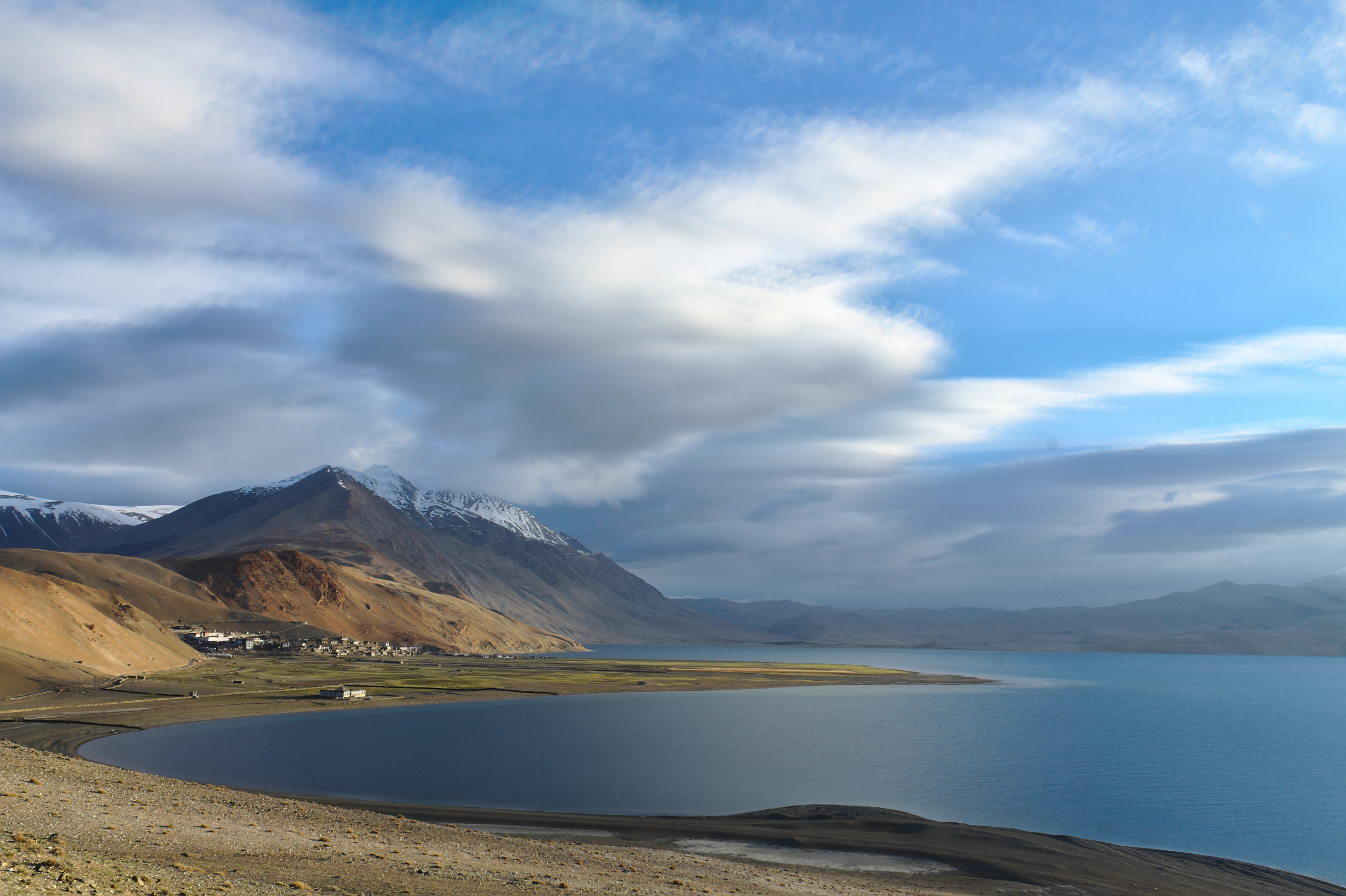 Una vista amplia del paisaje del lago Tso Moriri, que muestra agua azul profunda, una orilla rocosa de color tostado y laderas montañosas cubiertas de nieve bajo un cielo parcialmente nublado.
