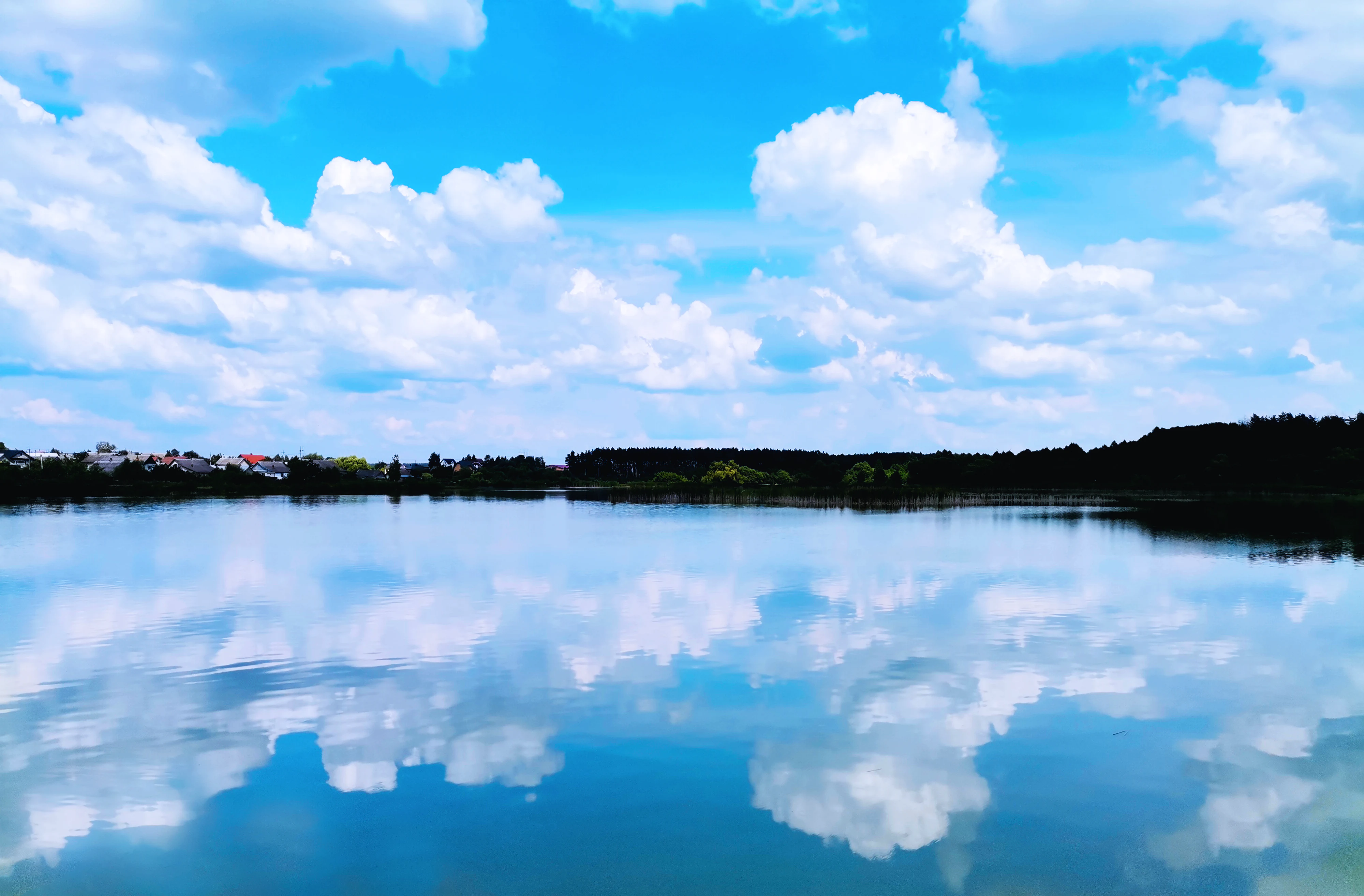 Calm pond reflecting clouds and sky with distant trees and small buildings on the horizon