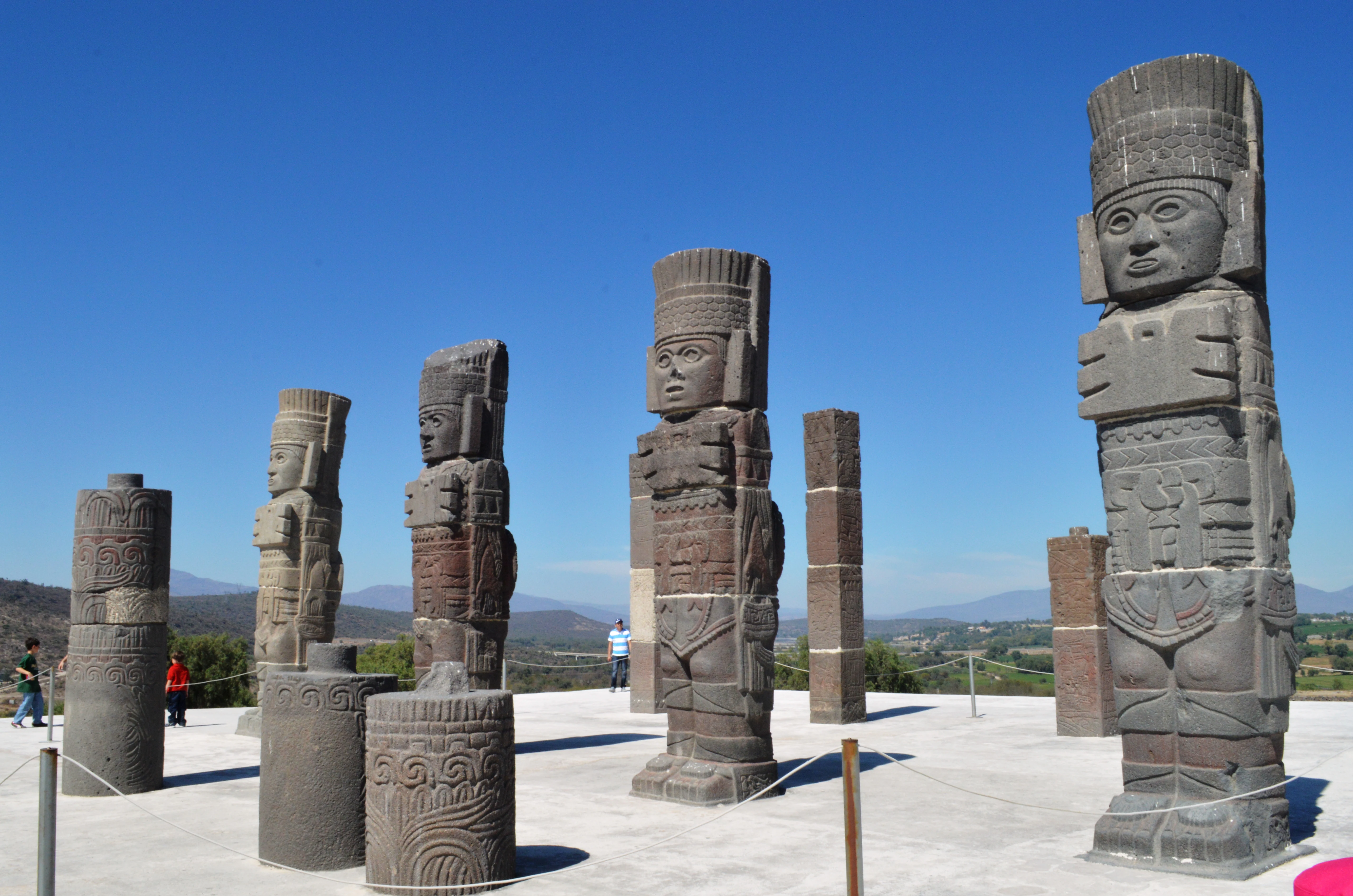Five large stone Atlantes statues on a paved platform with mountains in the background under a clear blue sky