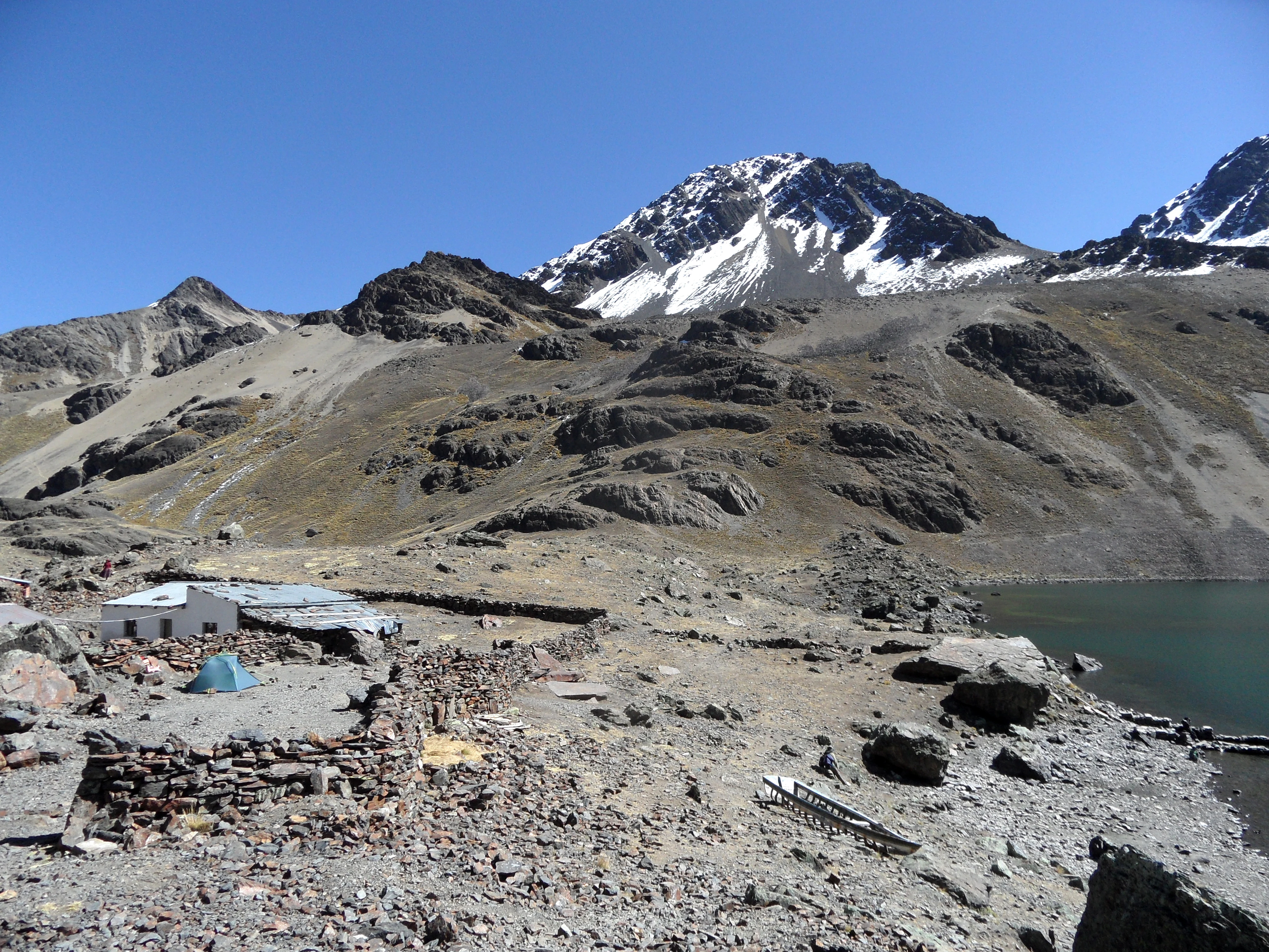 A rocky base camp area with a small stone structure and a blue tent near a glacial lake, set against a backdrop of snow-capped mountains in the Cordillera Real.