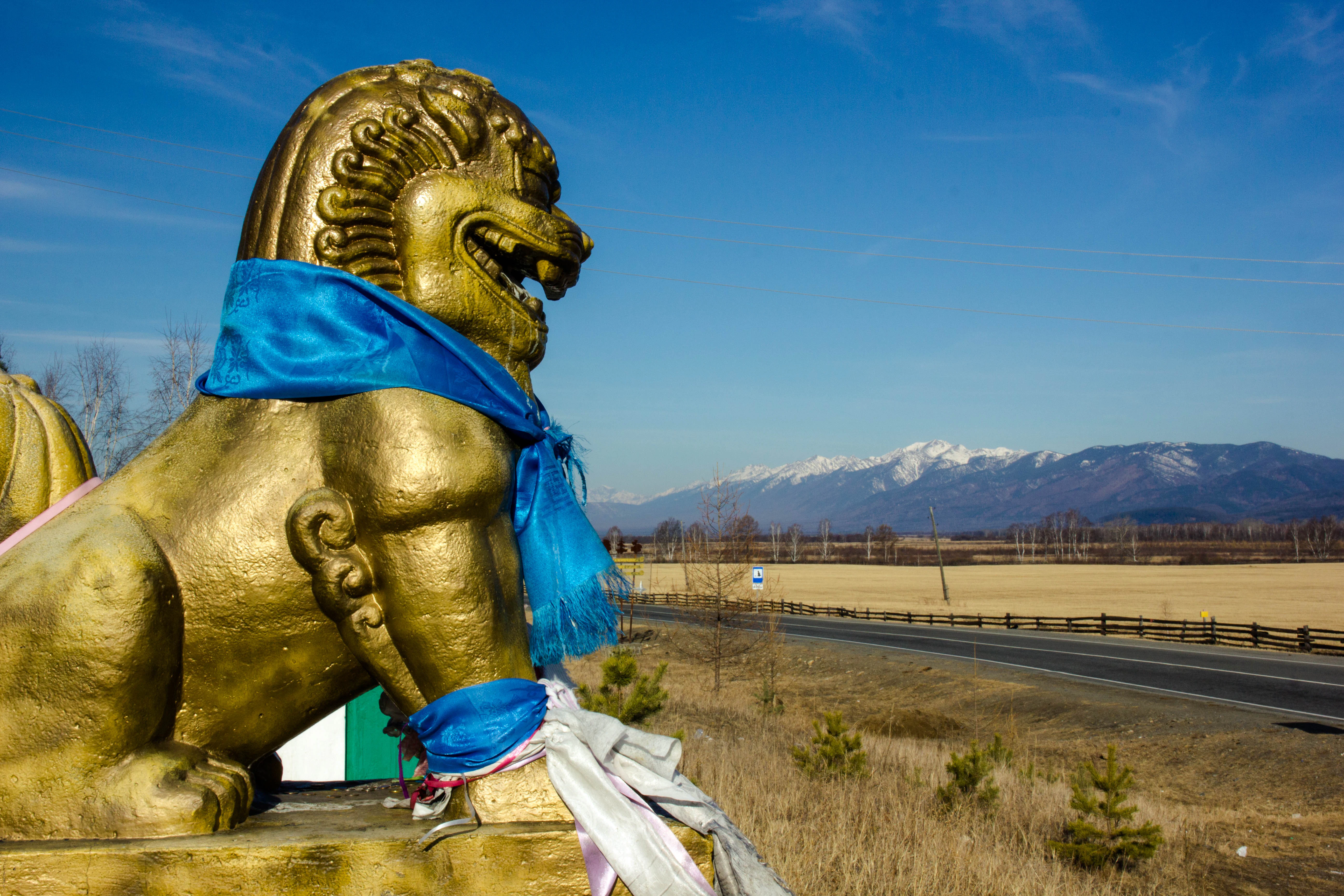 Golden lion statue adorned with blue scarves, set against a backdrop of distant mountains and a road under a clear blue sky