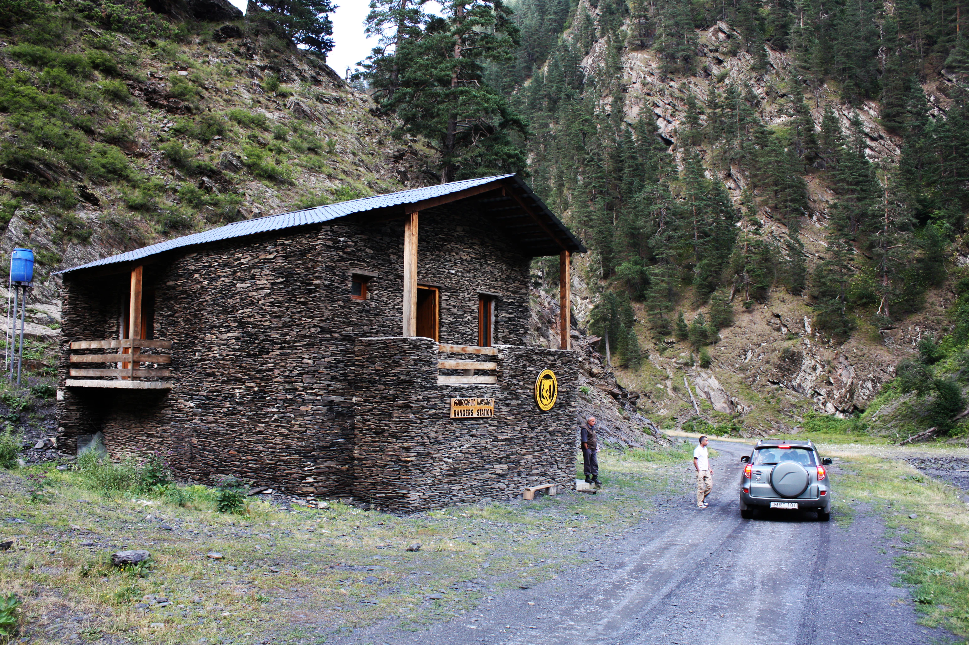 Stone building with signage, two people, and a vehicle on a gravel road in mountainous terrain.