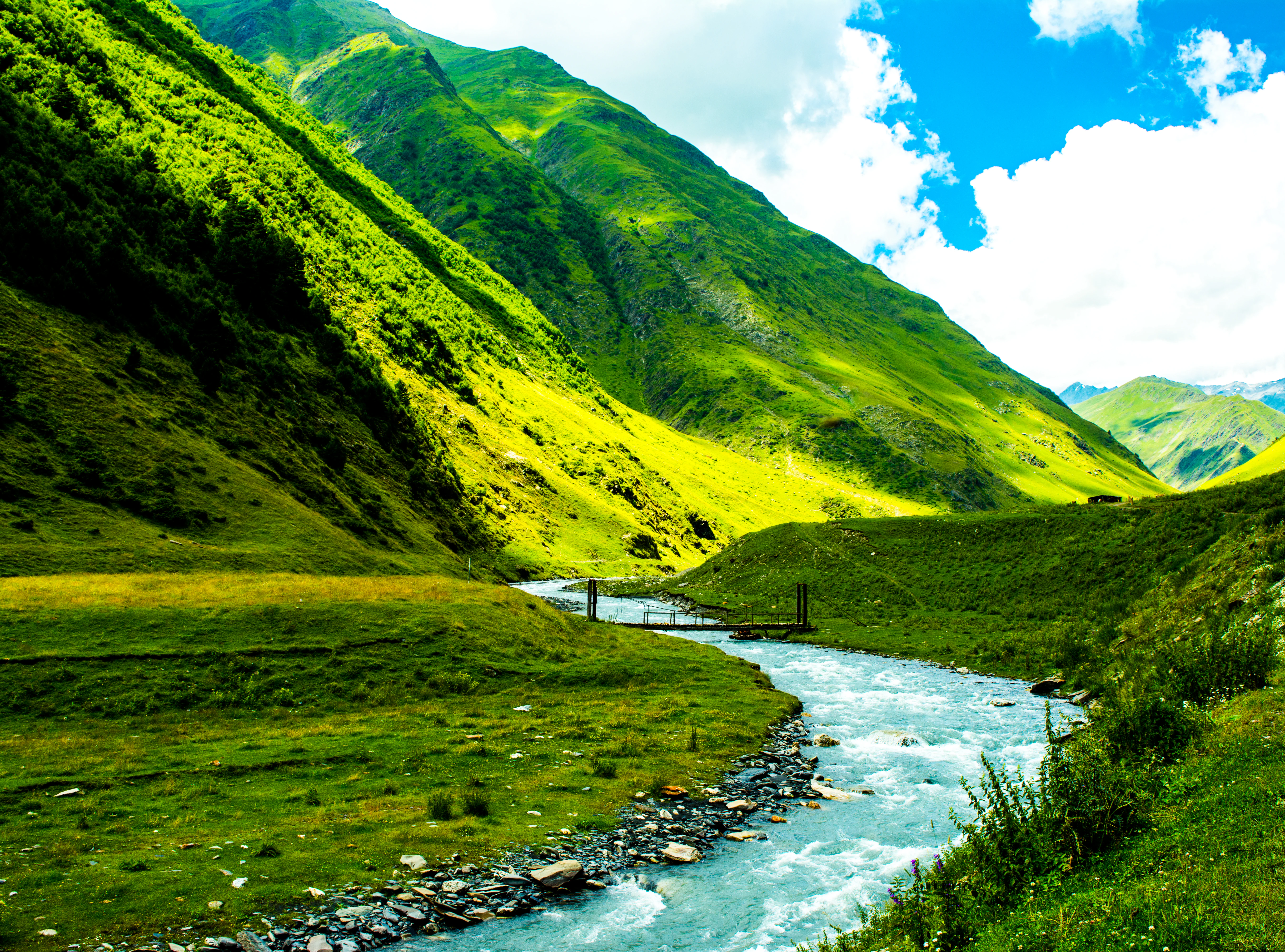 A river flows through a grassy valley with steep green mountains under a partly cloudy sky