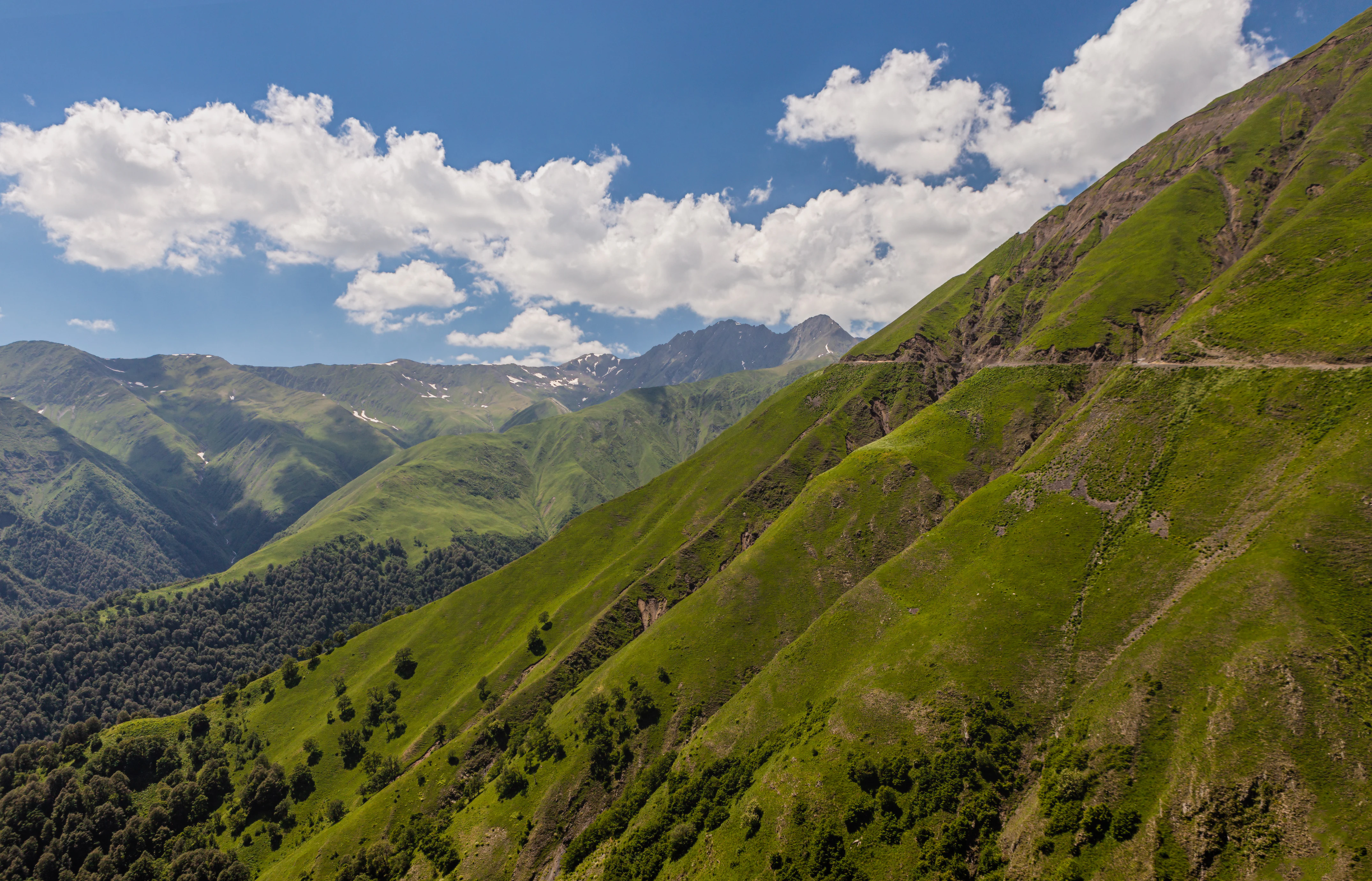 Green mountain slopes with a winding road under a blue sky with scattered clouds.