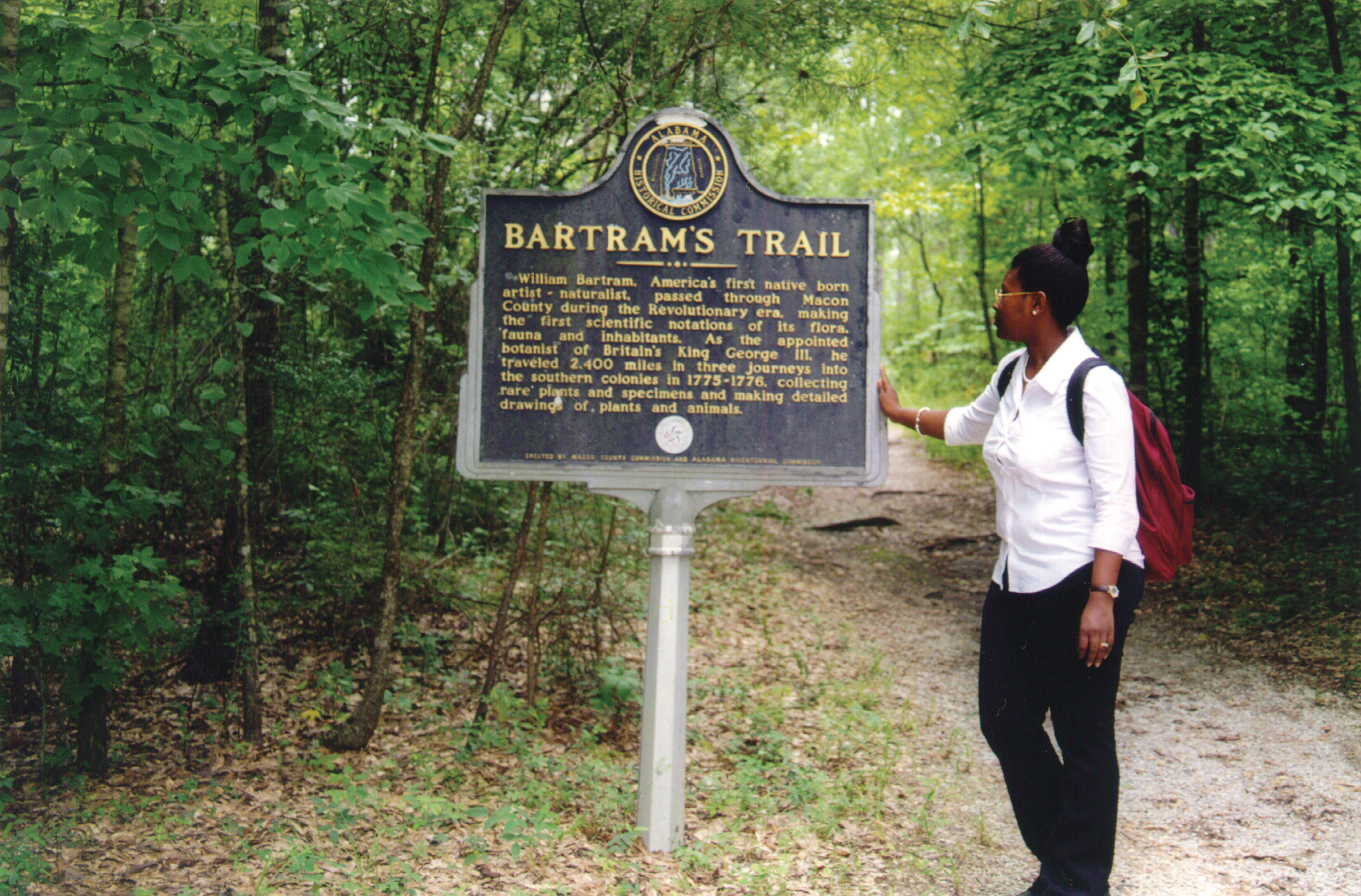 A woman stands on a dirt trail in a wooded area, reading a metal interpretive sign titled Bartram's Trail.