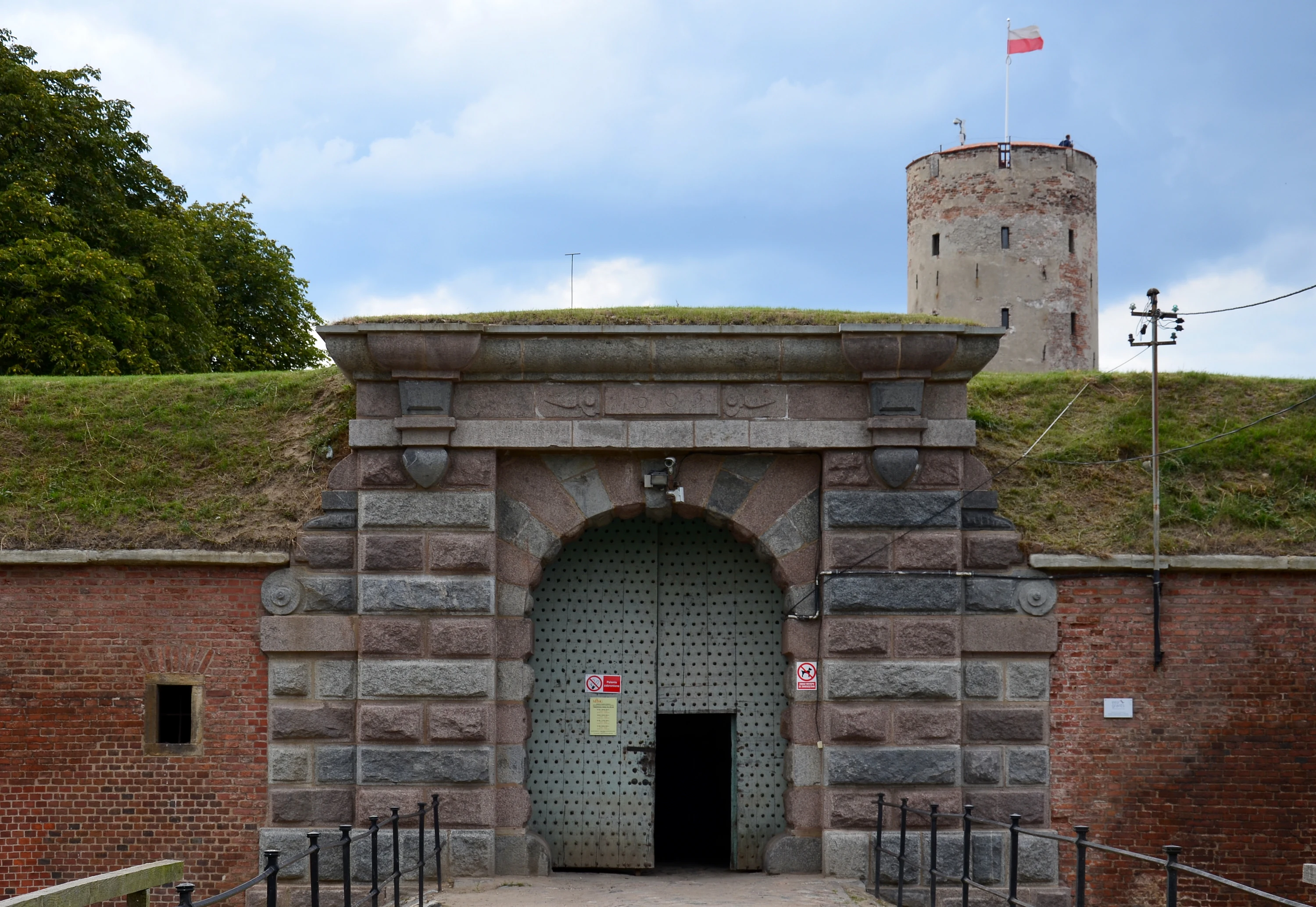 A stone archway with a large metal gate set into a brick and grass fortification, with a round brick tower flying a flag behind it.