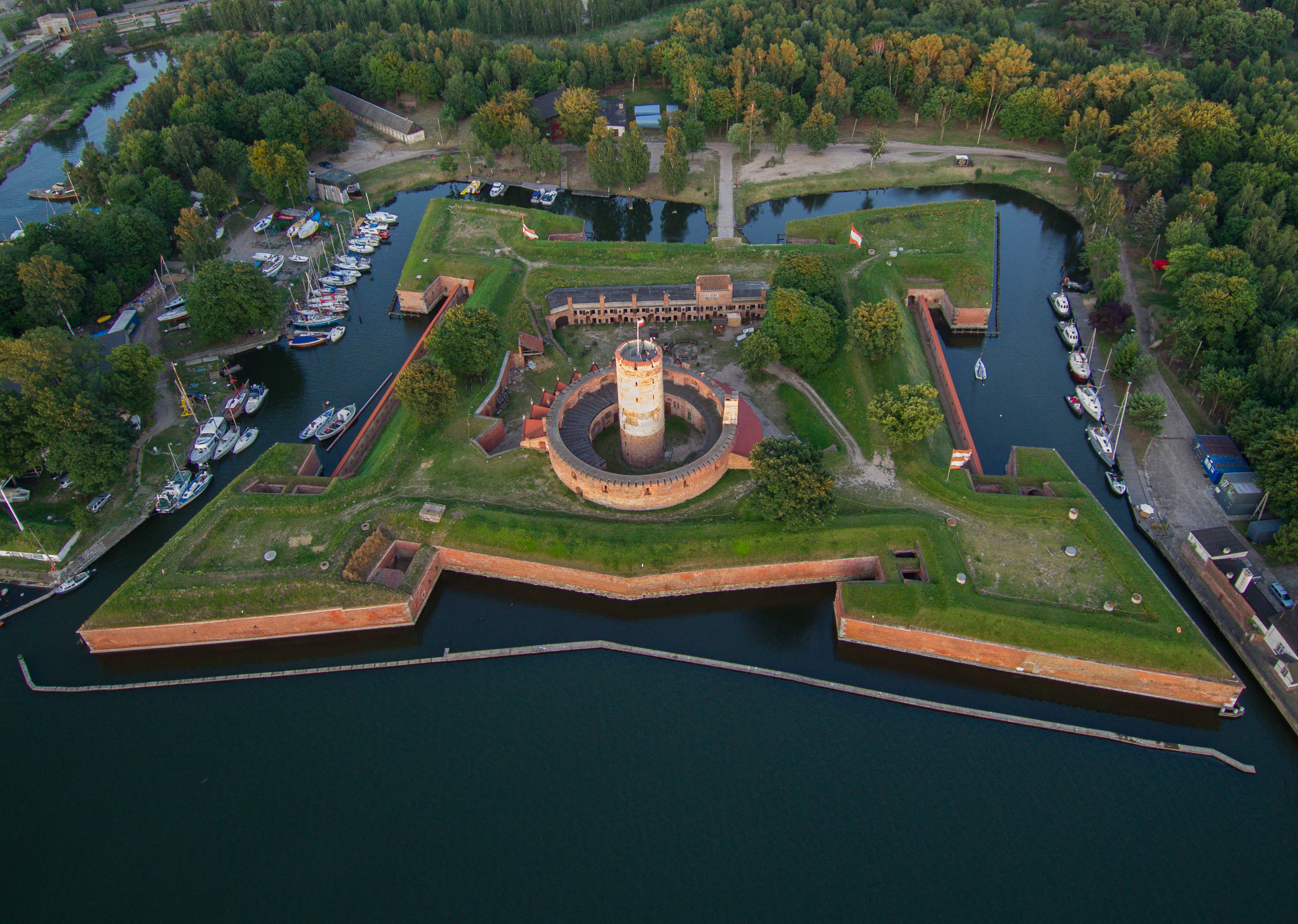 An aerial photograph of a star-shaped brick fortress with a central round tower, surrounded by dark water, with boats docked along the inner canals and green trees in the background.