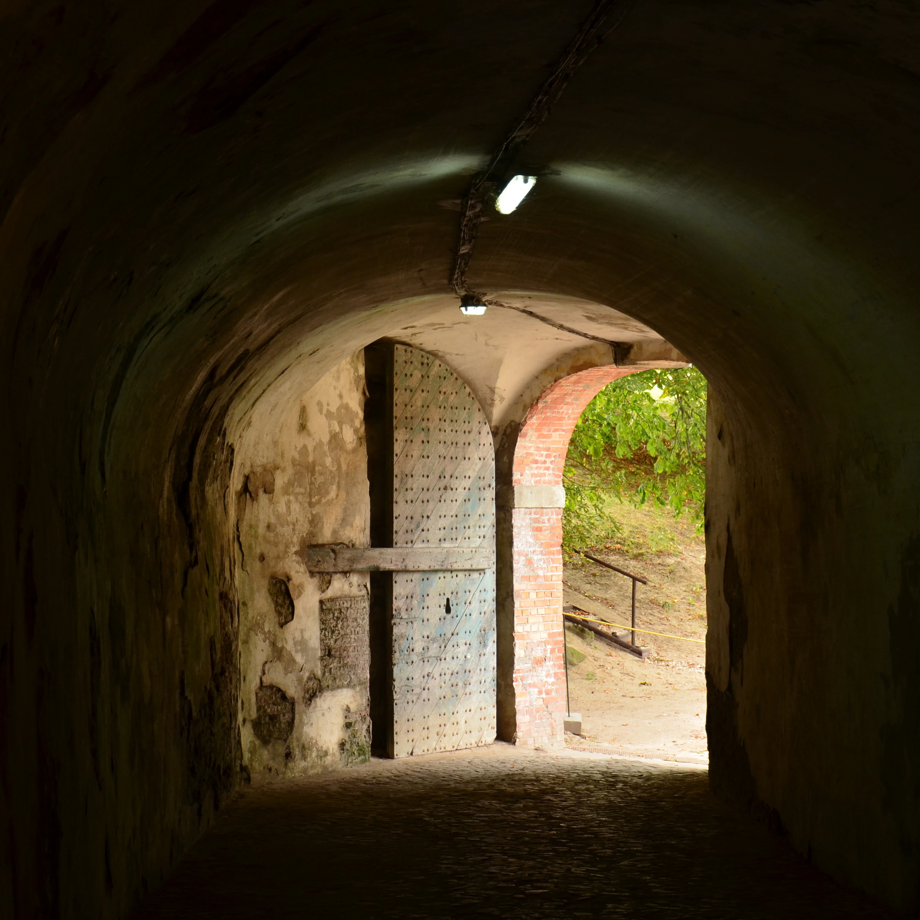 Dark arched stone tunnel with a large door on the left and an opening showing green vegetation outside.