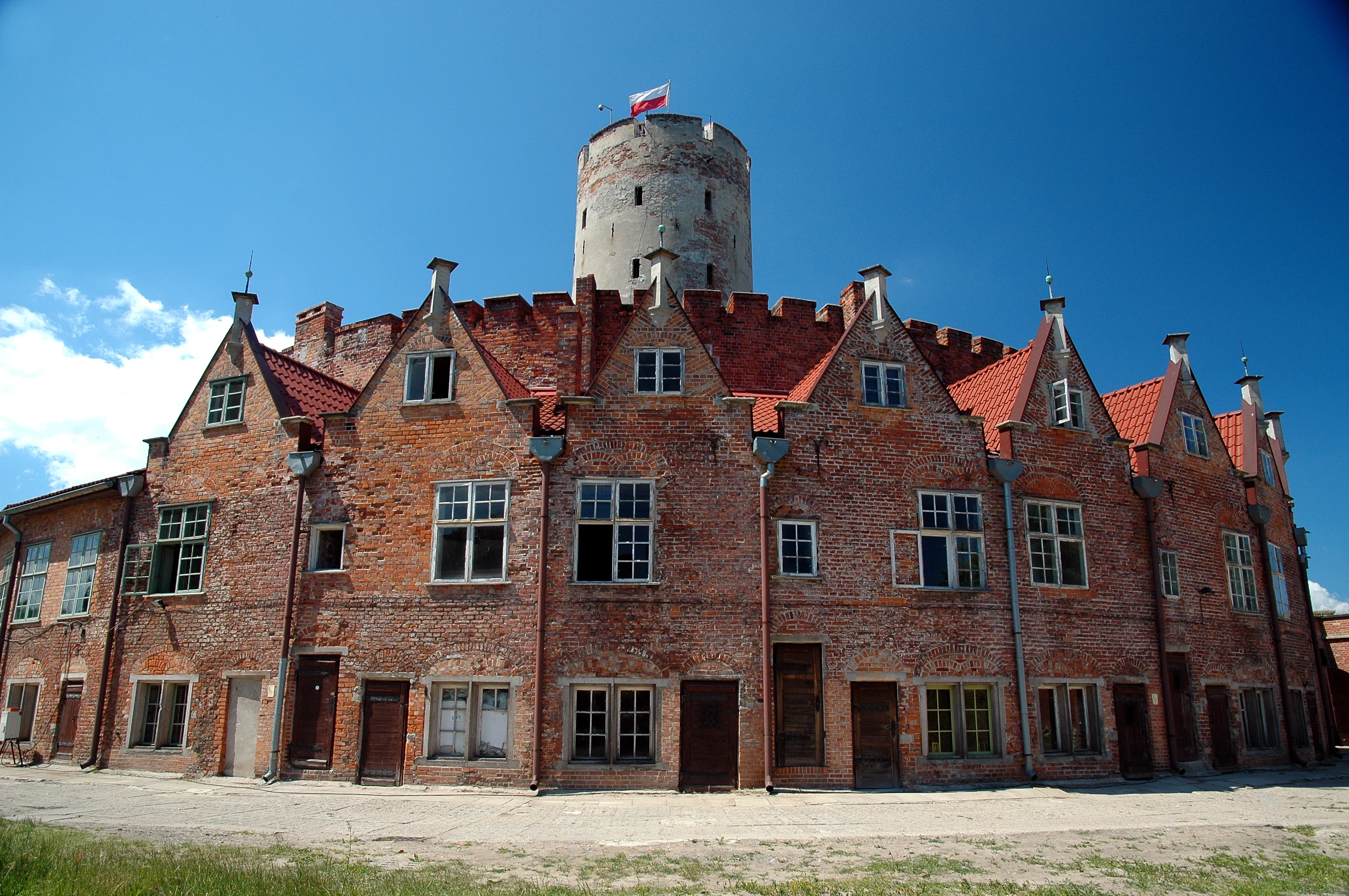 A wide-angle shot of a large, curved red brick building with multiple gabled roofs and a tall cylindrical stone tower rising from the center, topped with a flag, set against a blue sky.