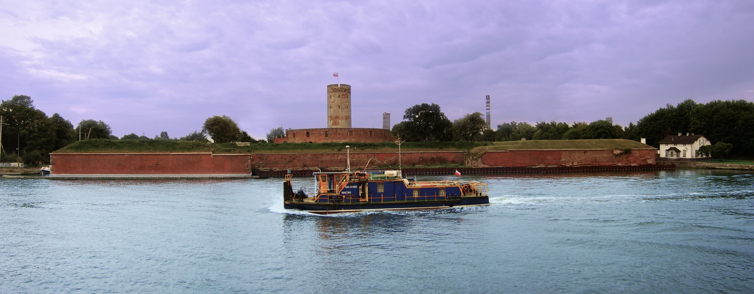 A wide panoramic photograph showing a large red brick fortress with a central tower situated on a riverbank under a cloudy sky, with a blue and black boat moving across the water in the foreground.