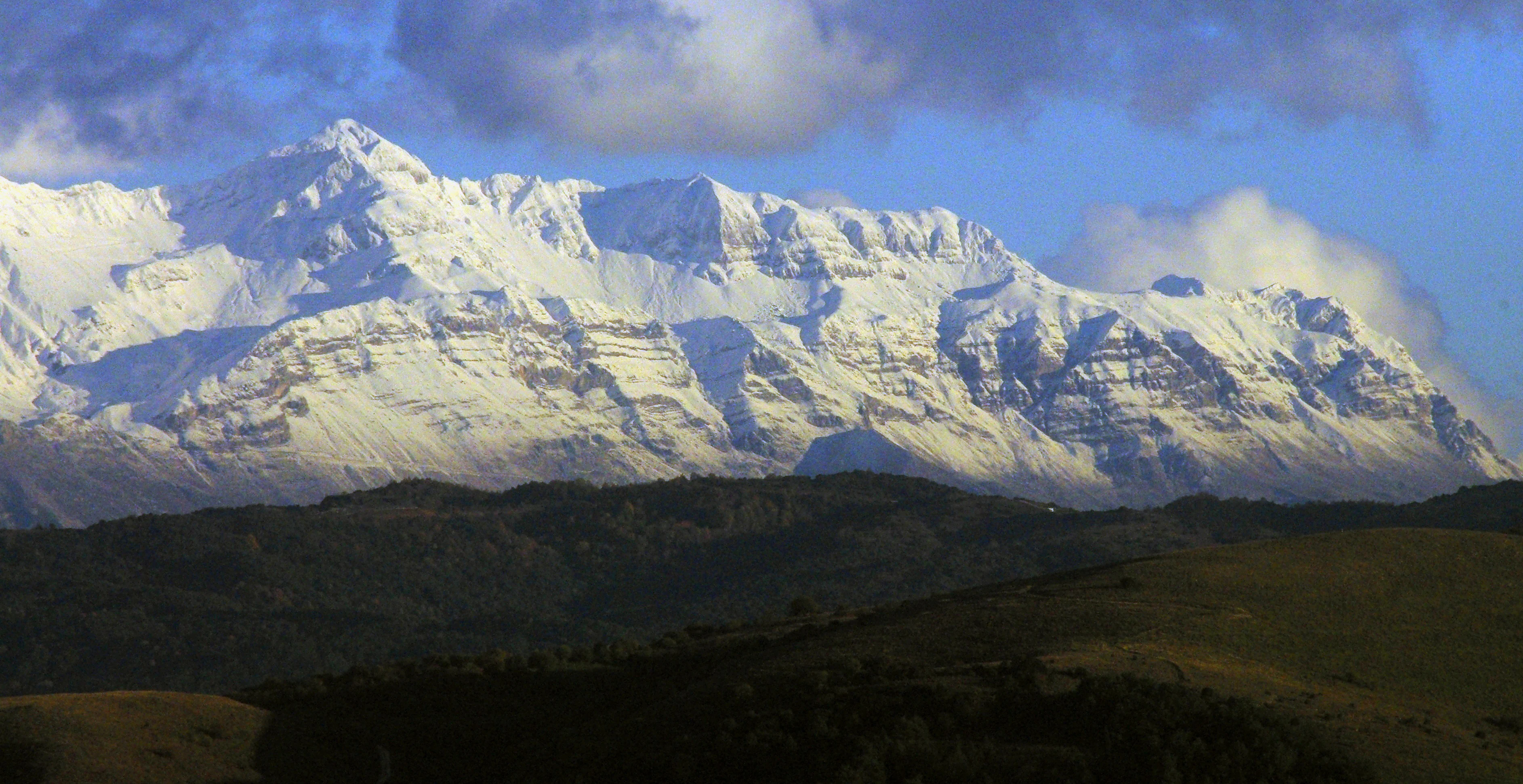 Panoramic view of a jagged, snow-covered mountain range towering above dark green foothills under a partially cloudy blue sky.