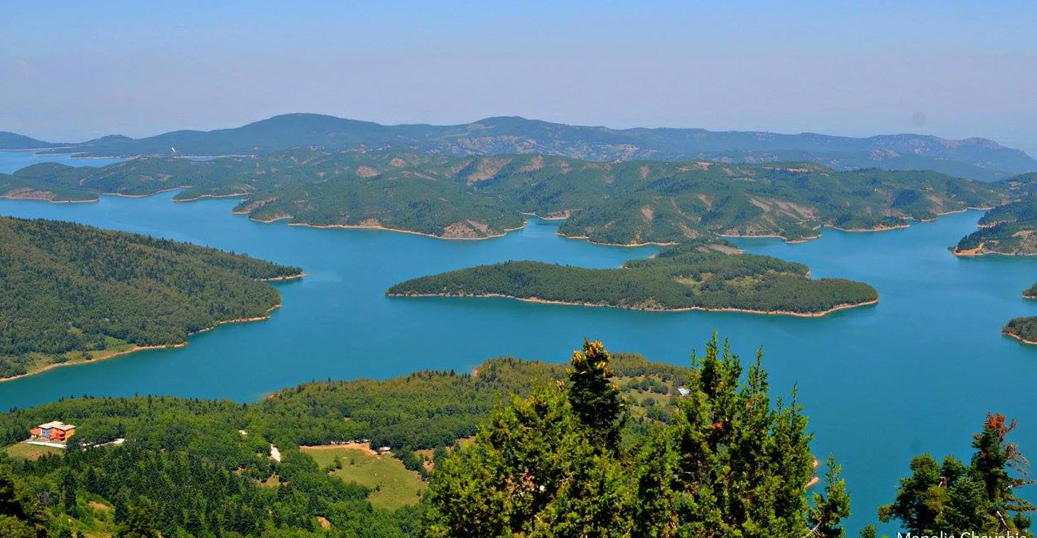 Panoramic landscape view overlooking a large, calm, turquoise lake with several tree-covered islands and green mountainous shores under a clear sky.