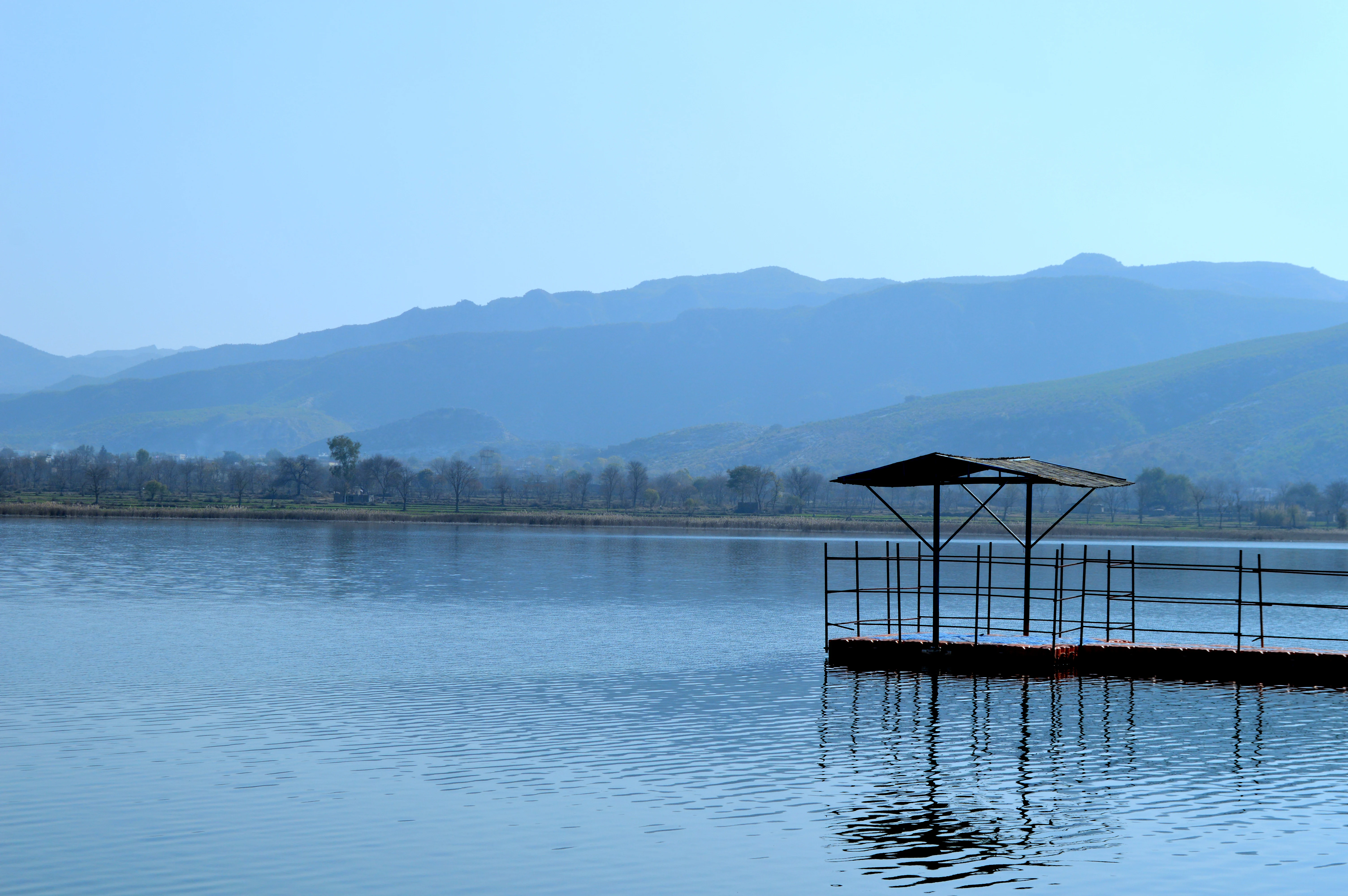 A calm lake with a small wooden pier featuring a roofed shelter, set against a backdrop of distant mountains and trees under a clear blue sky.