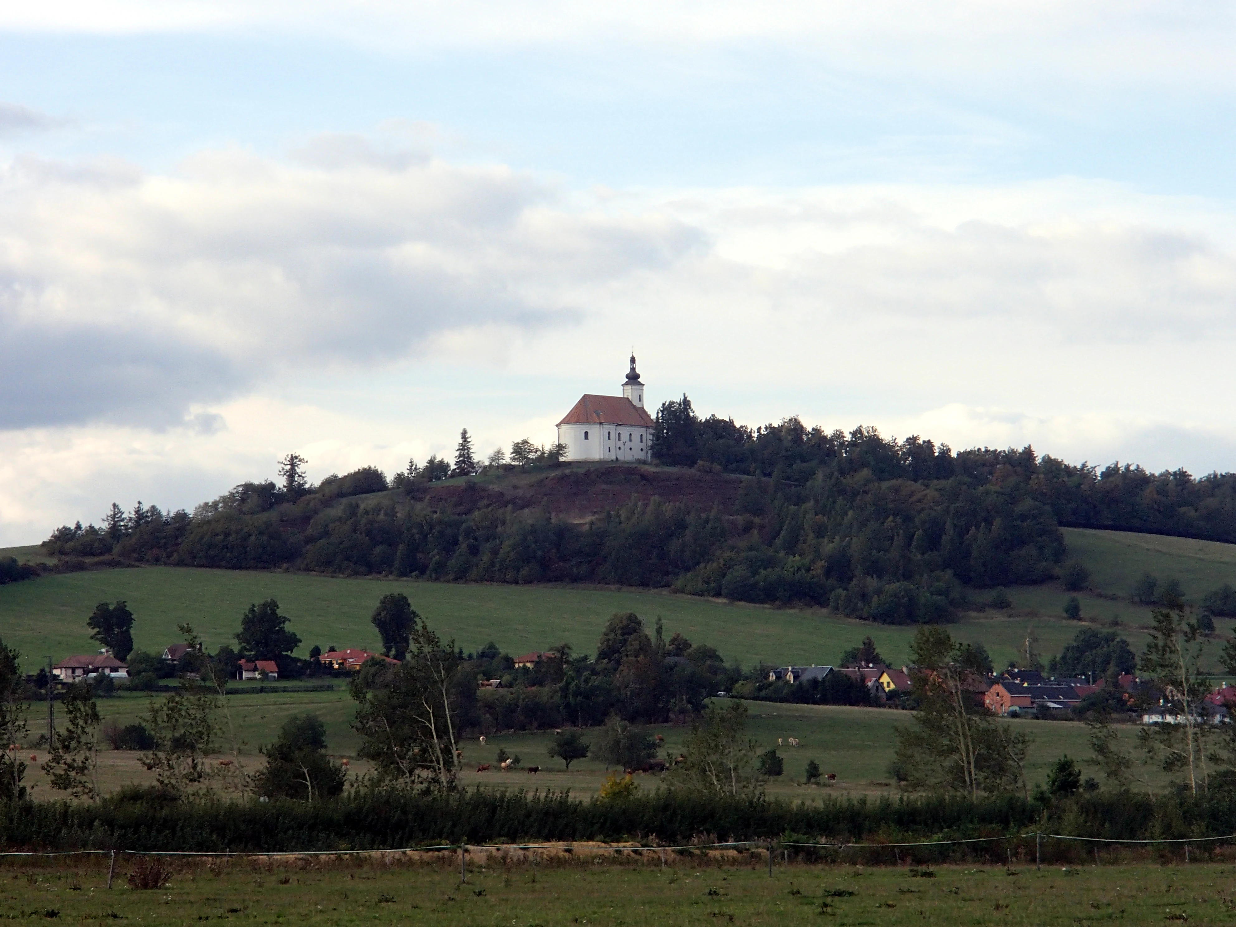 A wide landscape view of a green hill topped with a white pilgrimage church, surrounded by trees and a rural village at the base.