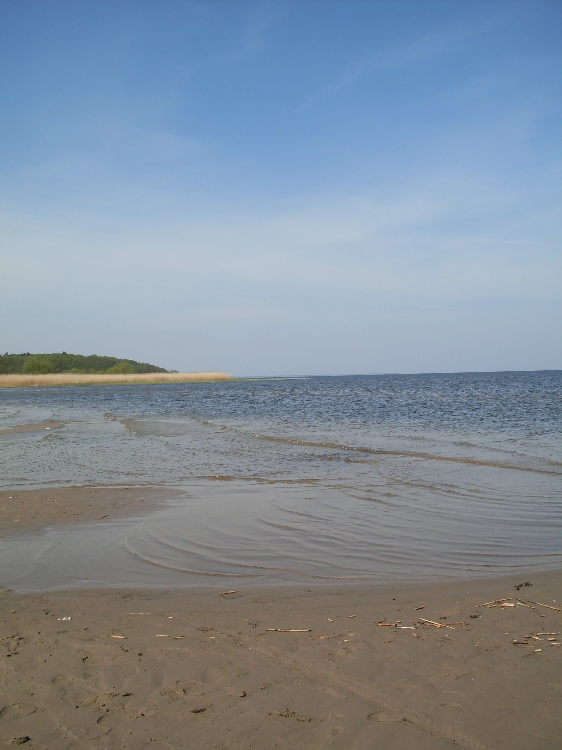 A sandy shoreline leading into a large, calm lagoon under a clear blue sky, with a reed-lined coast visible in the distance.