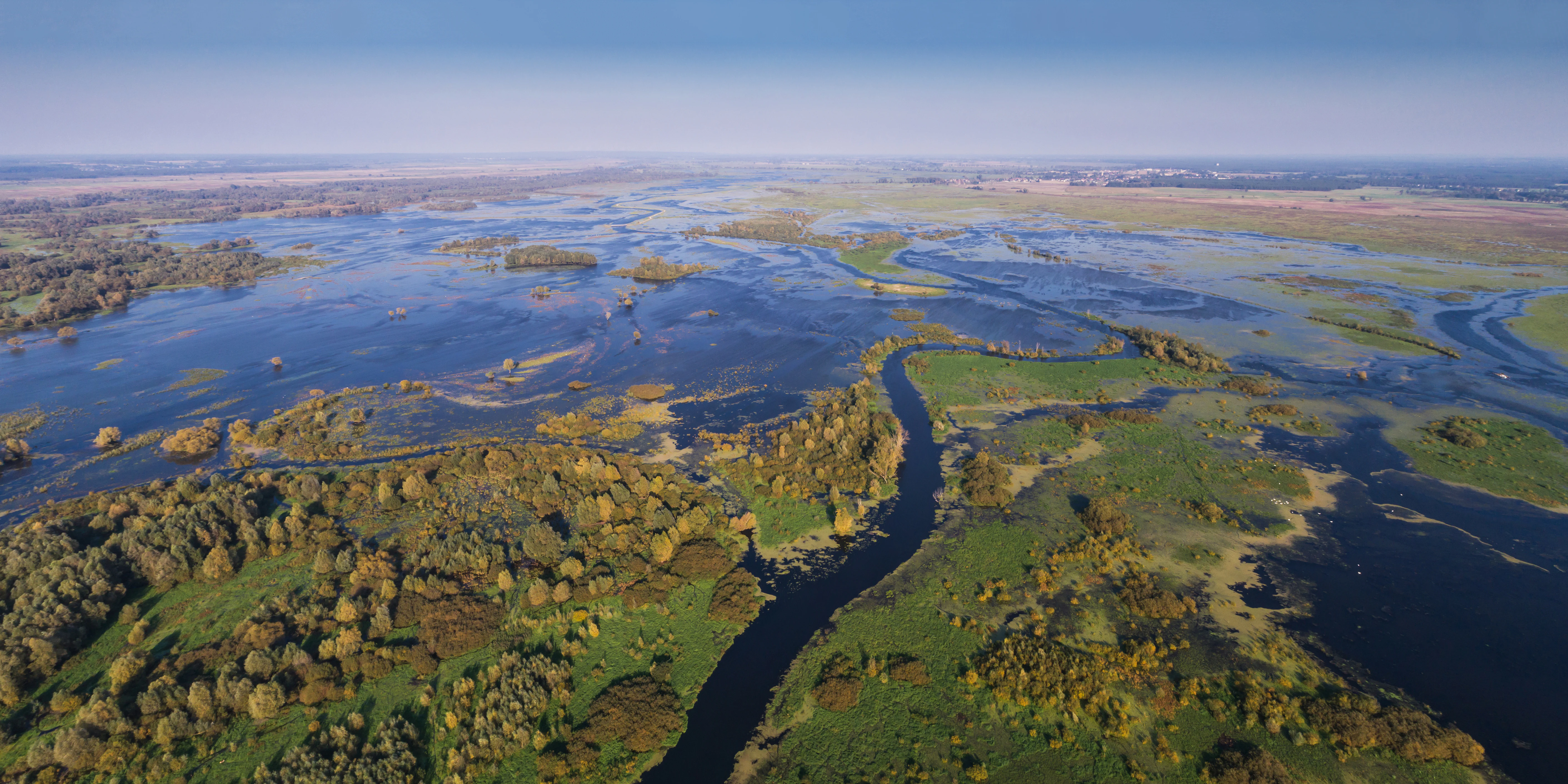 A wide aerial view showing extensive flooded marshlands and winding river channels meandering through lush green and autumnal forest patches.