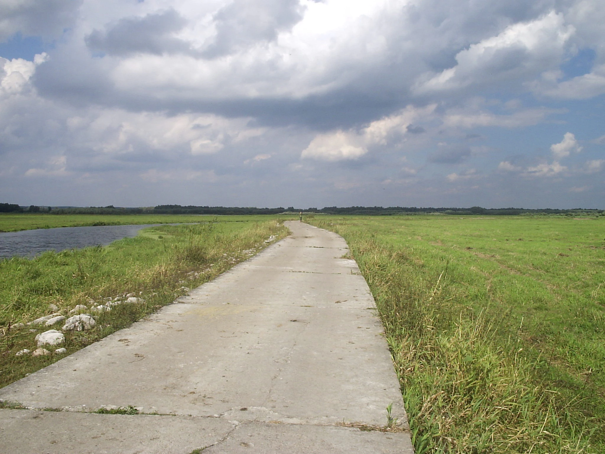 A narrow, worn concrete road stretches into the distance, flanked by lush green grasslands and a small river or waterway on the left under a cloudy sky.