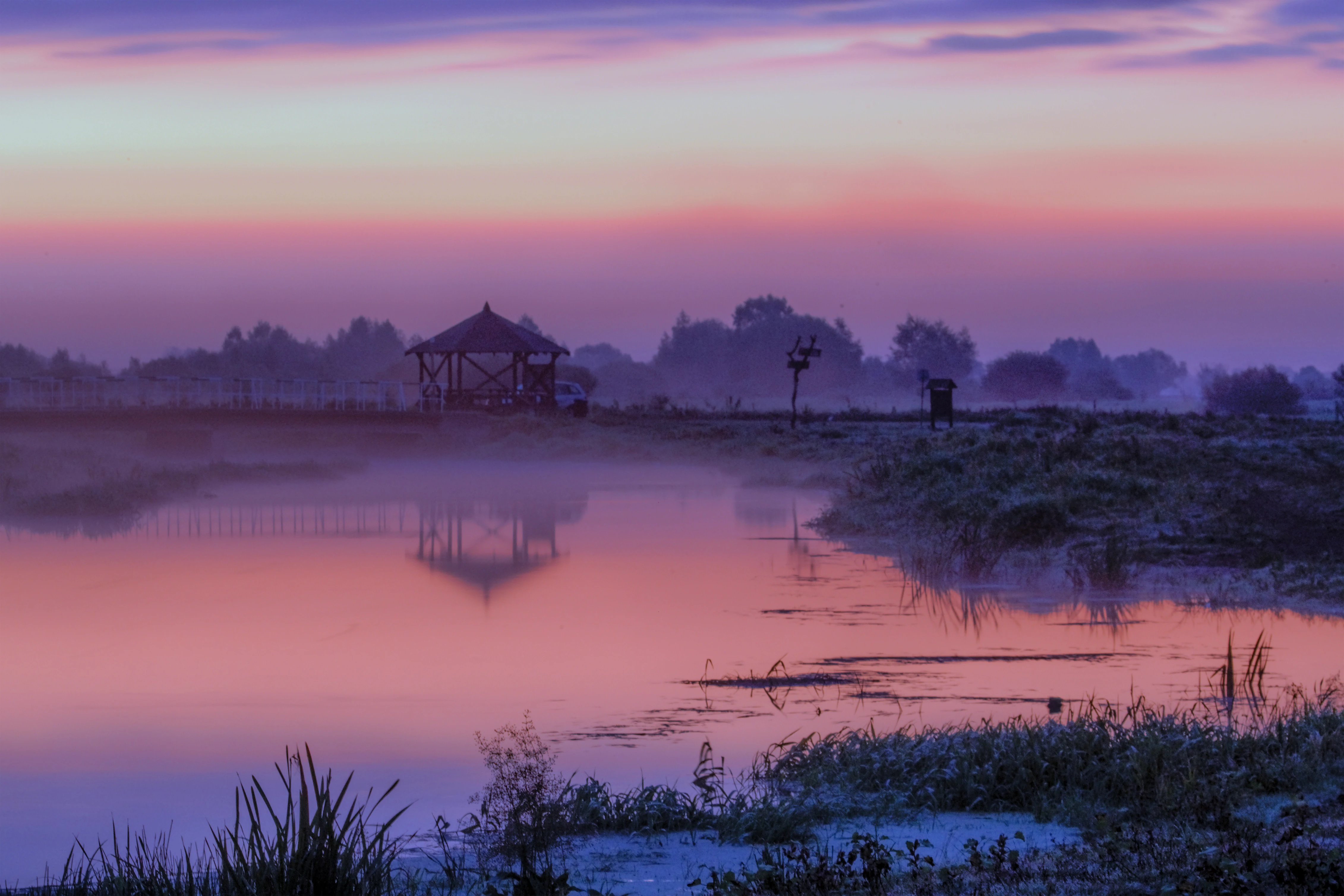 A peaceful sunrise scene over a still river and wetland area, with a wooden bird observation shelter silhouetted against a purple and orange sky.