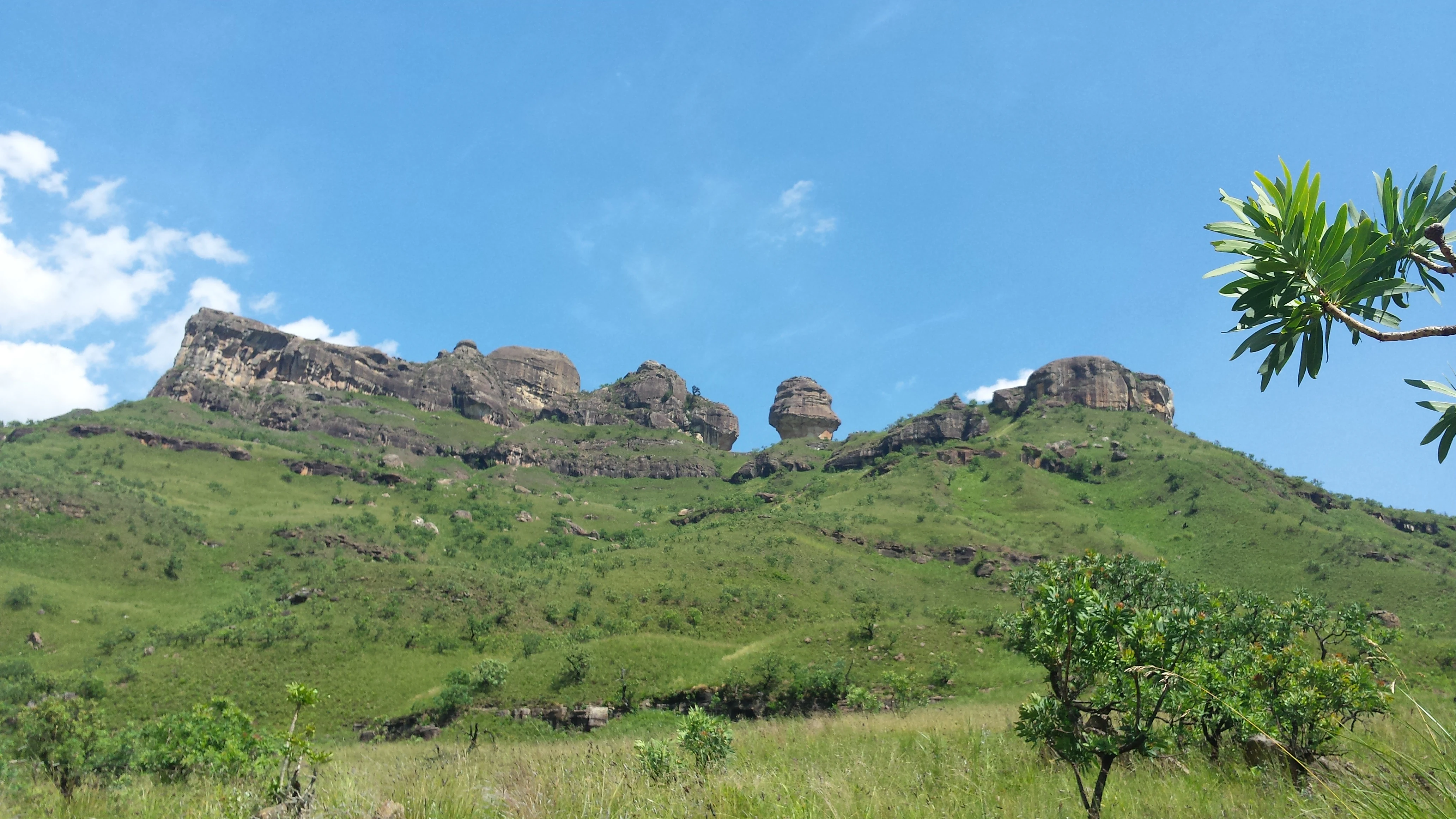 A green, grassy mountainside leading up to a rugged cliff band under a clear blue sky, with a prominent standalone rock formation known as Policeman's Helmet.