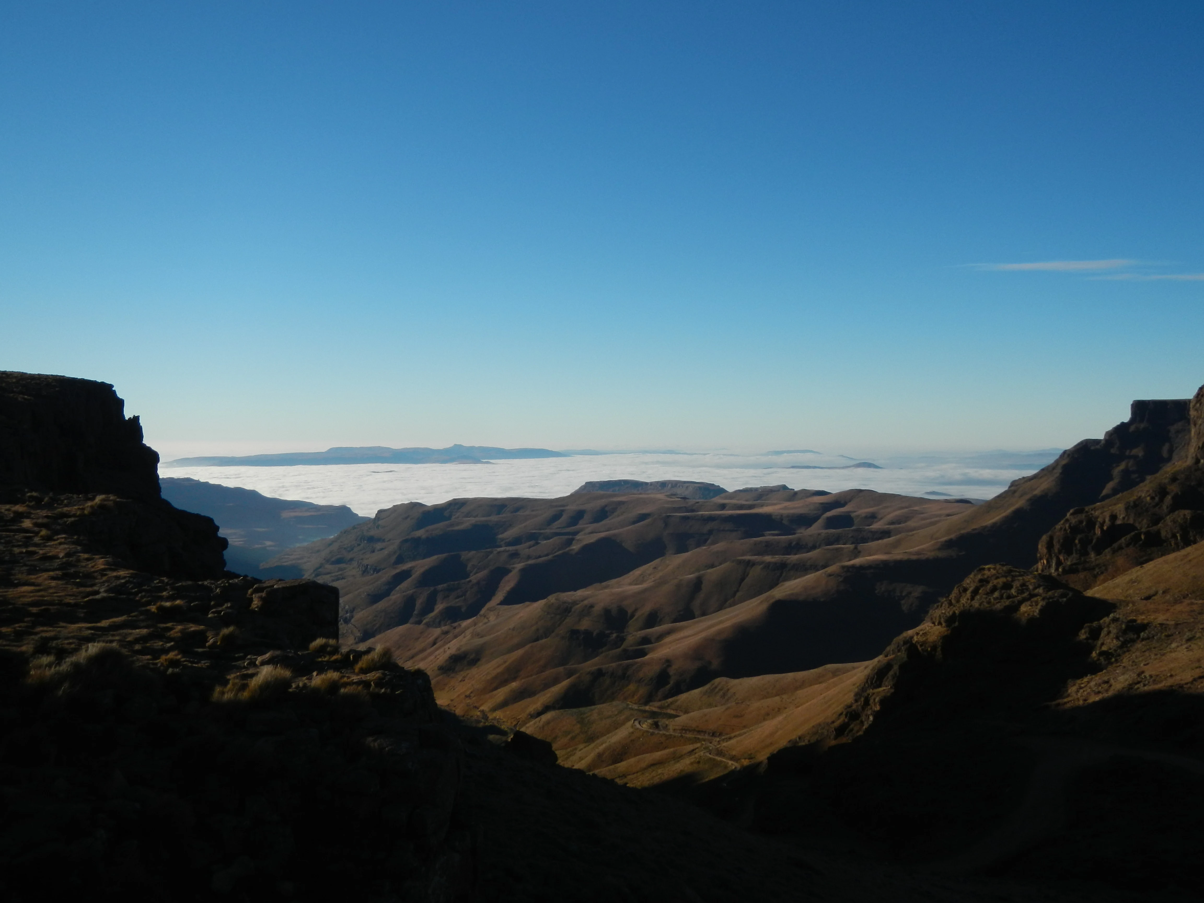 A wide view from a high cliff edge looking over rolling, shadowed mountain slopes toward a distant, low-lying cloud bank under a clear blue sky.