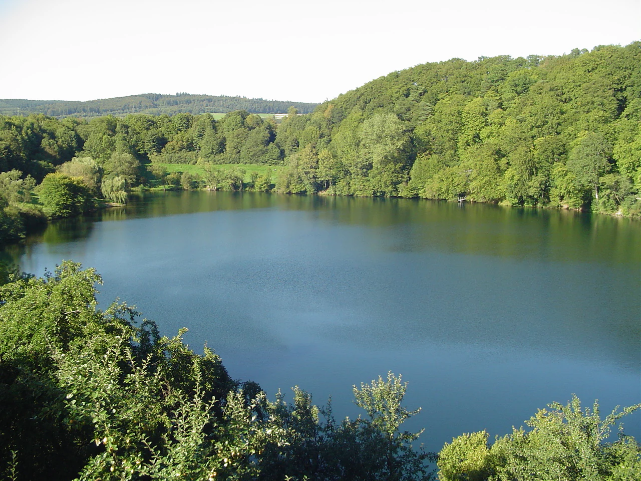 A high-angle view of a calm, dark blue circular lake surrounded by lush green forested slopes and a distant rolling horizon.