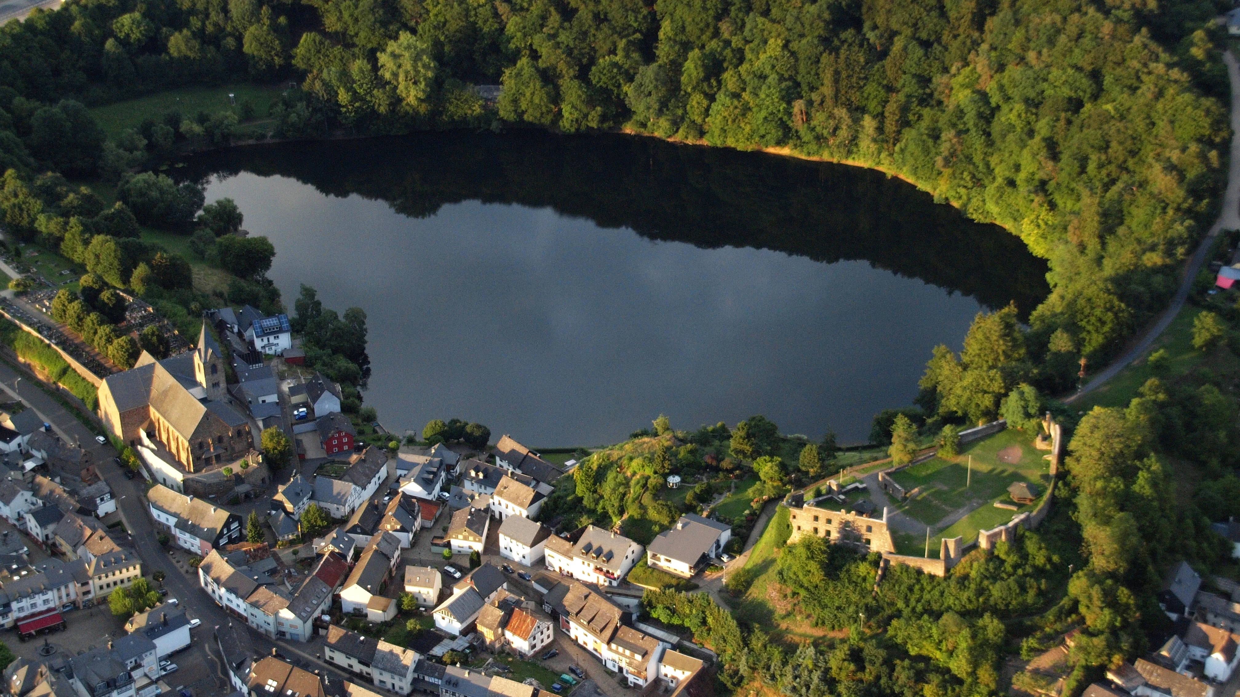 An aerial perspective of a dark, circular volcanic crater lake surrounded by dense forest, with the town of Ulmen and castle ruins visible along the shoreline.