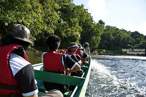 Grupo de personas con chalecos salvavidas rojos a bordo de un bote largo en un río con una densa selva tropical de fondo.