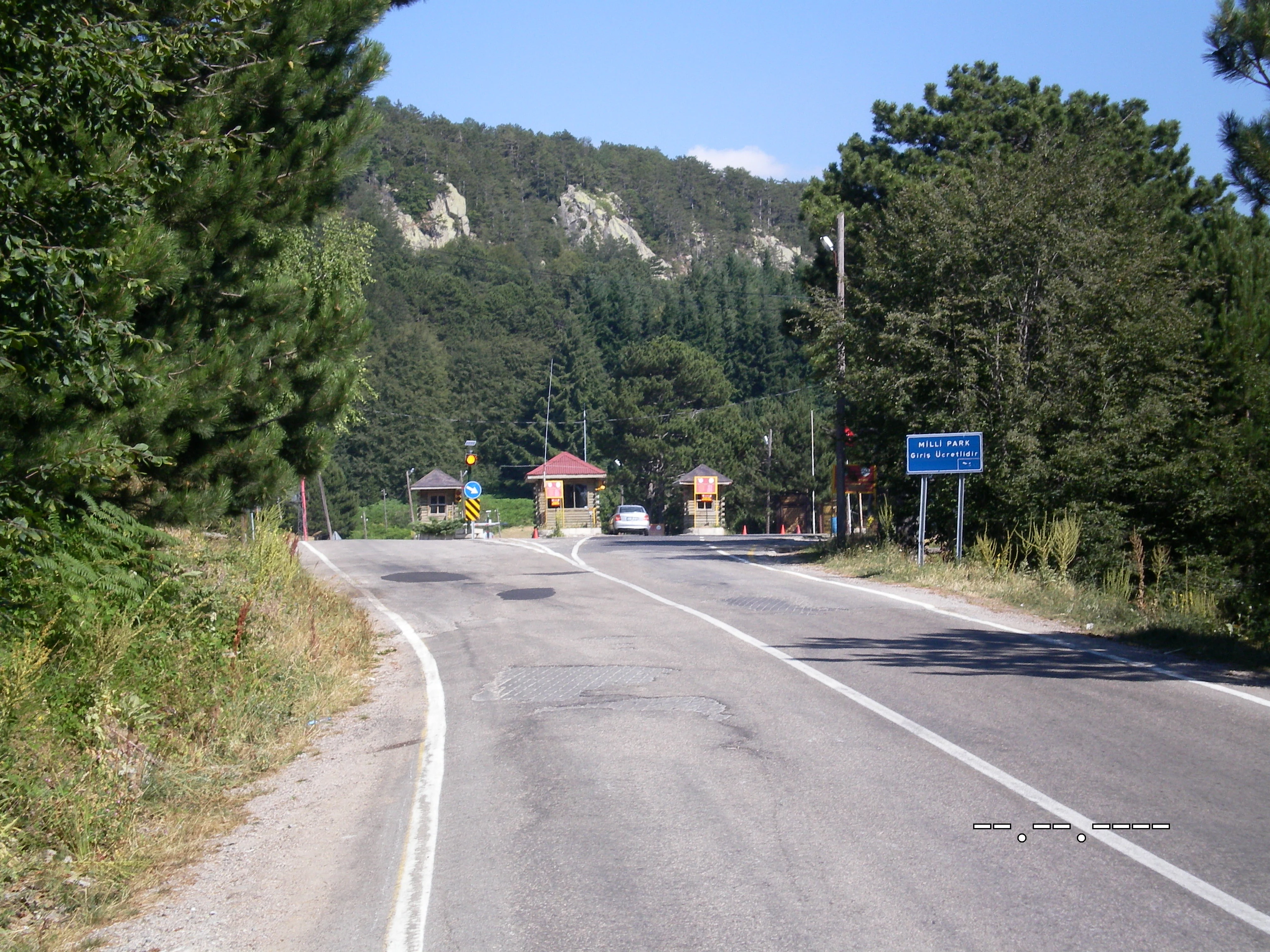 Paved road leading to Uludağ National Park entrance flanked by trees with blue signboard and gate buildings visible