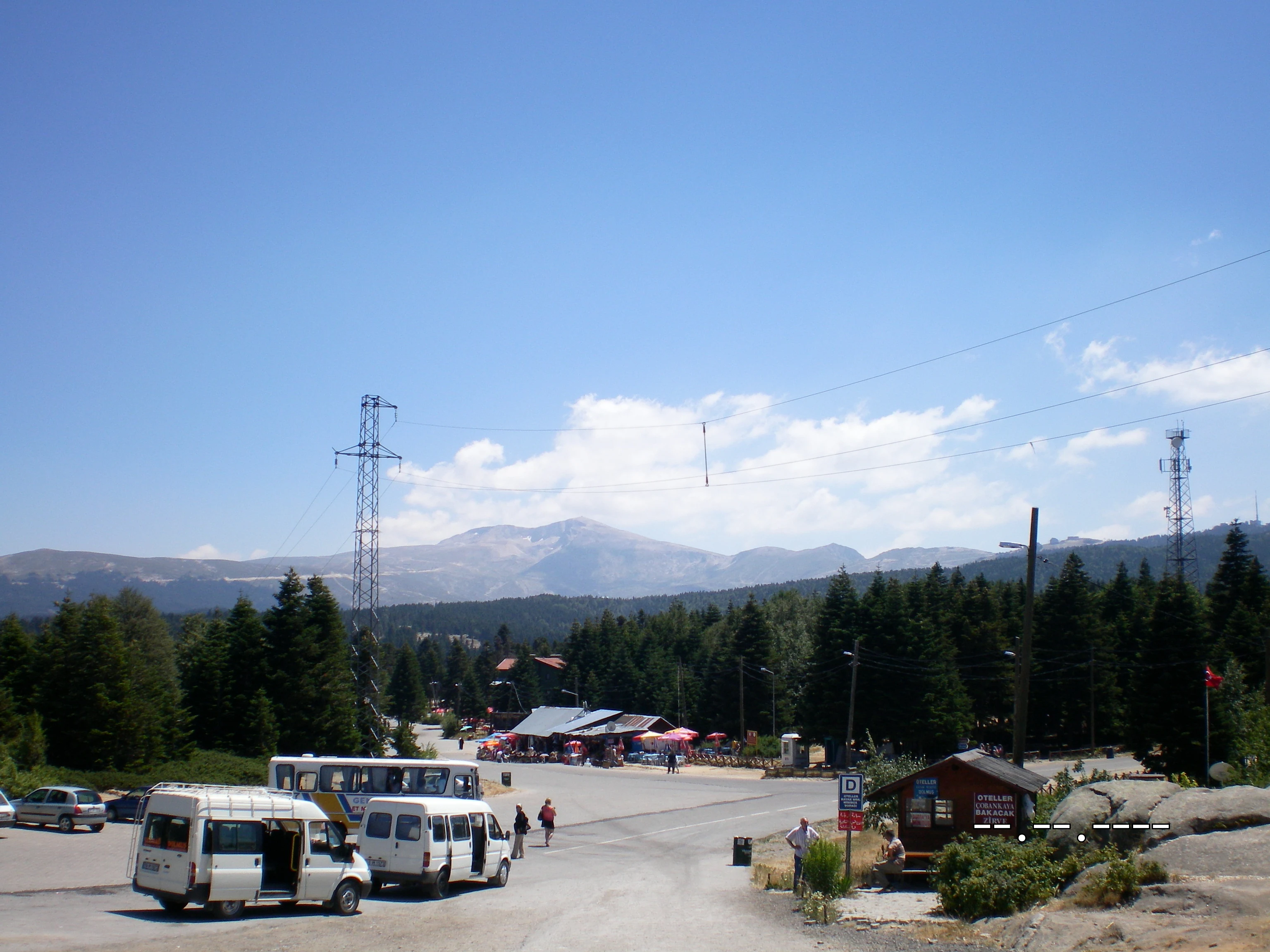 View of Uludağ National Park with white vehicles on a road, small building, forested hills, and mountain peaks under a clear blue sky