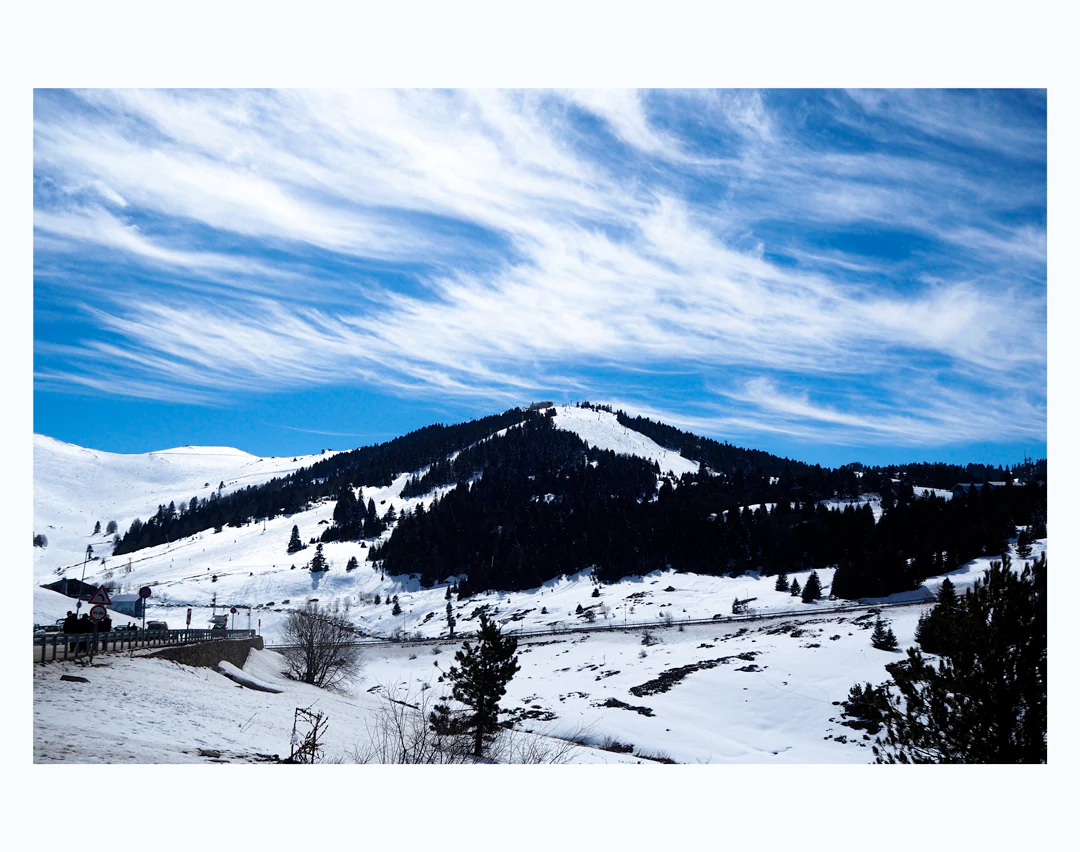 Snow-covered mountain slopes with a winding road, scattered trees, and a blue sky with wispy clouds