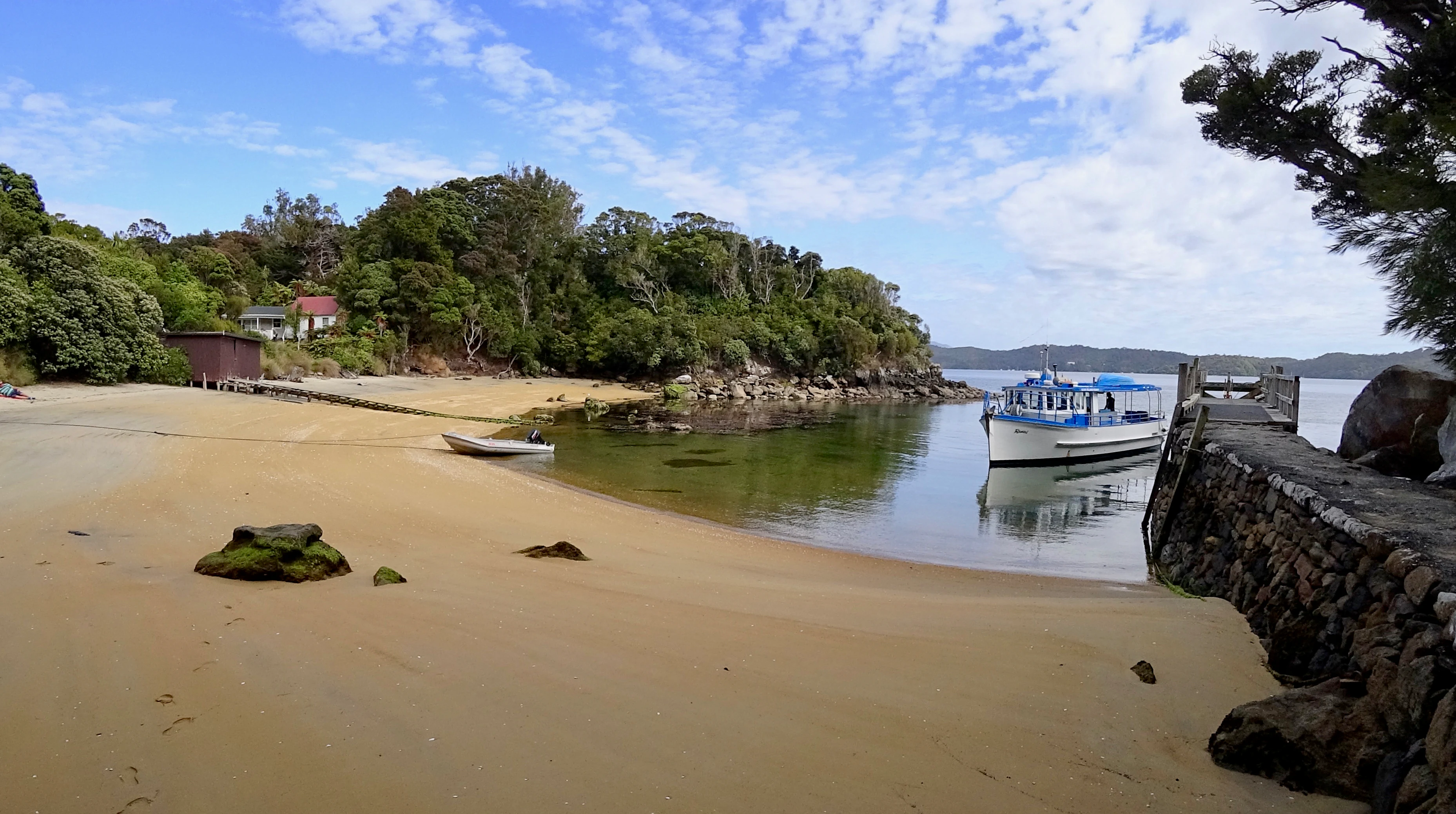 A sandy beach and stone wharf at Post Office Bay on Ulva Island, with a small passenger ferry anchored nearby and dense forest in the background.