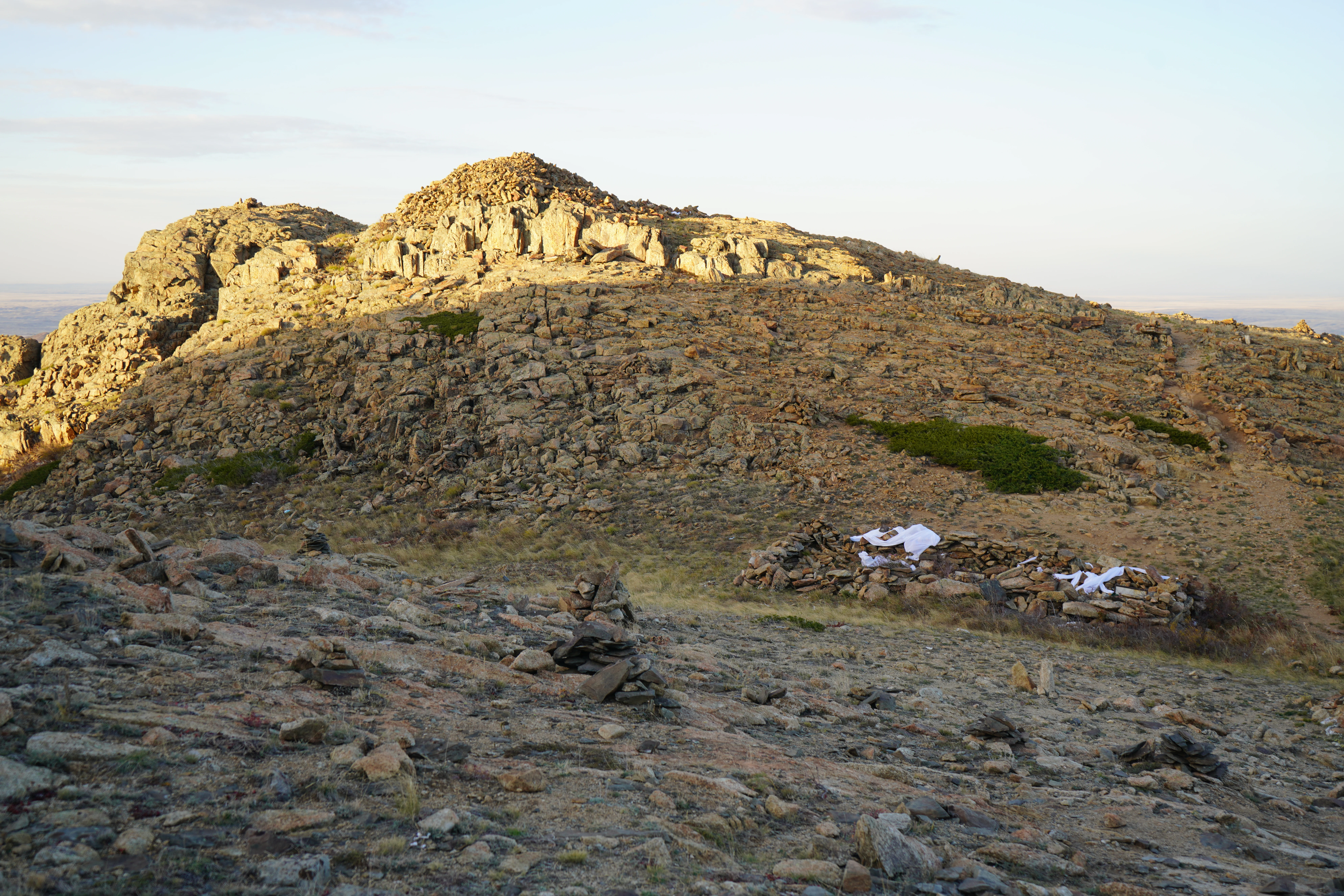 A rocky, arid mountain peak under soft, warm light in Ulytau National Park, featuring a small stone structure on the slope.