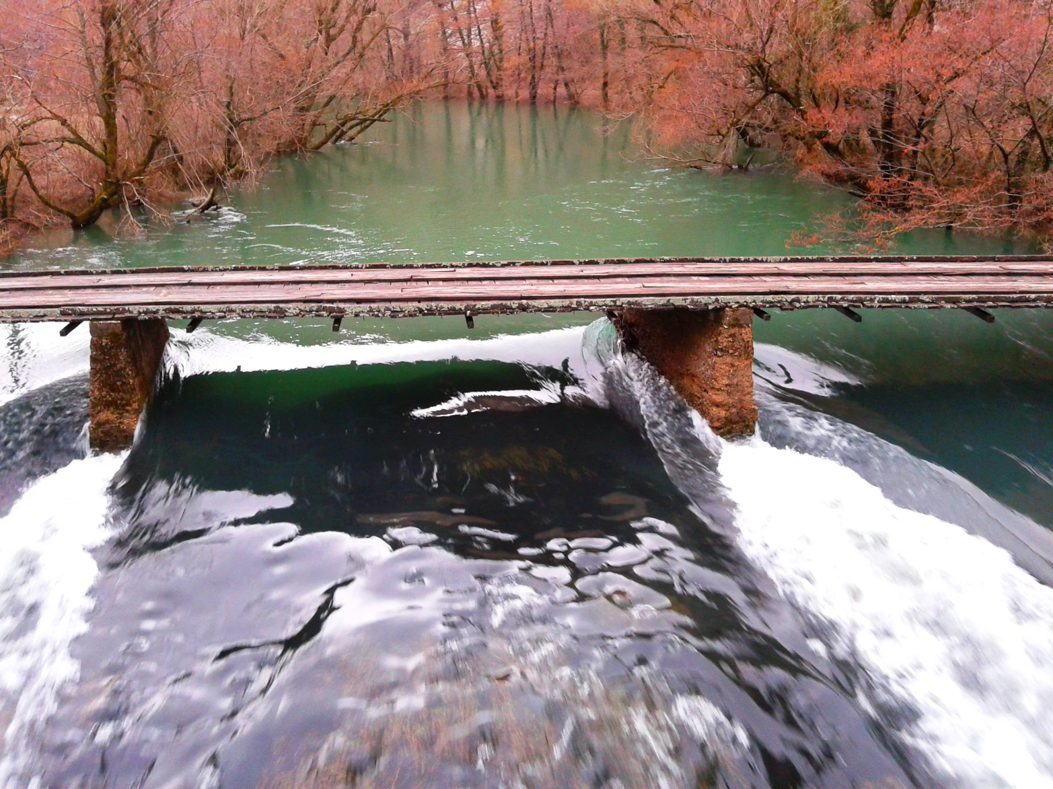 A wooden bridge with stone pillars crossing a river with white waterfalls, surrounded by trees with autumn foliage