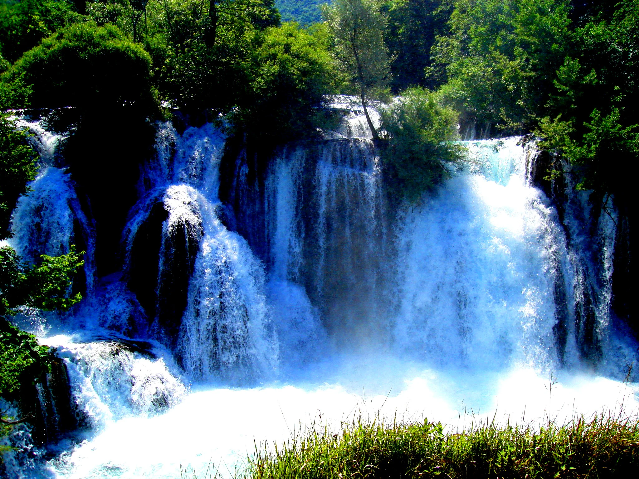 Multi-tiered waterfall flowing over rocks with lush green trees and vegetation surrounding it