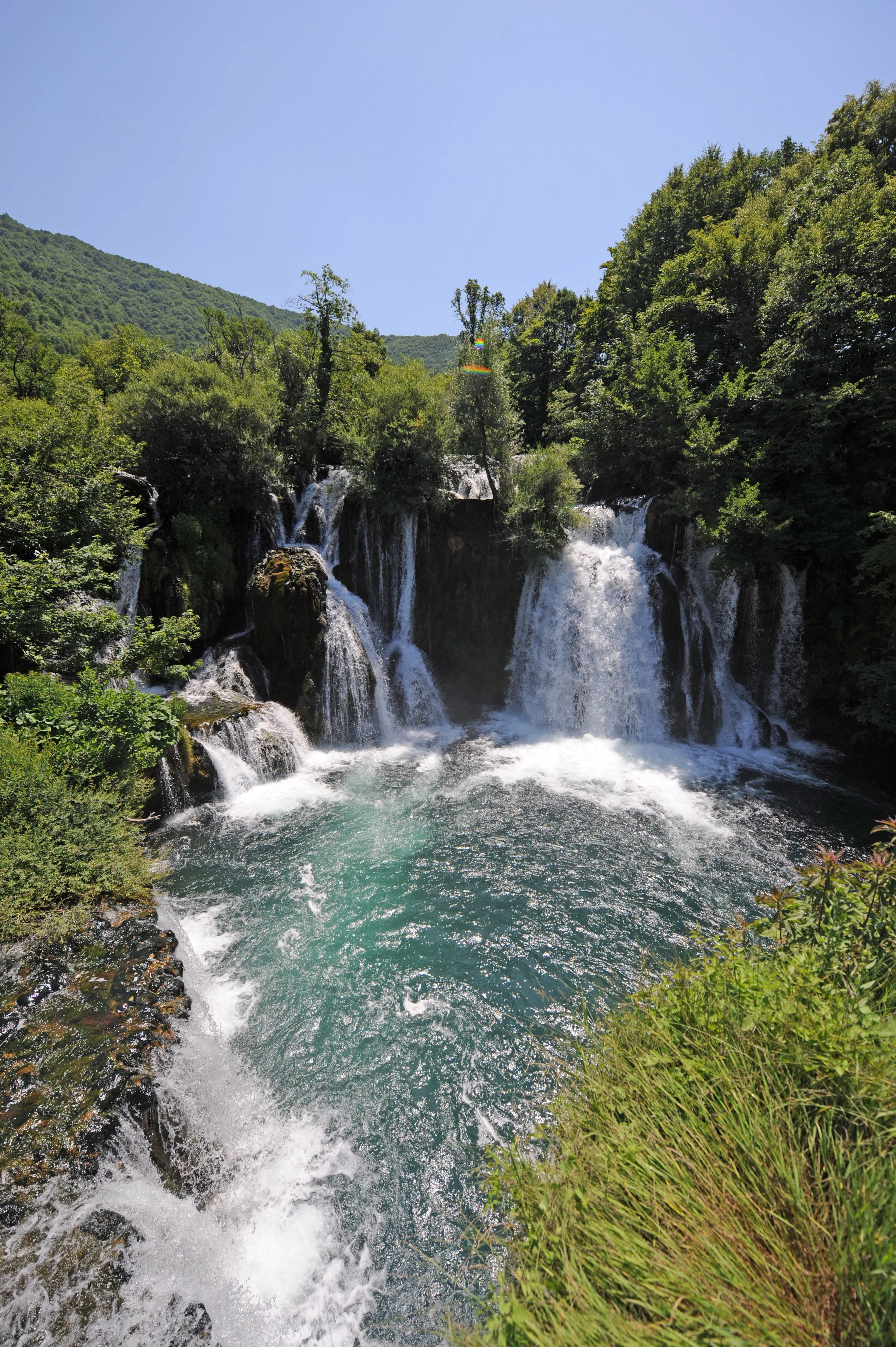 Multi-tiered waterfall cascading into turquoise pool surrounded by dense green vegetation under clear blue sky.