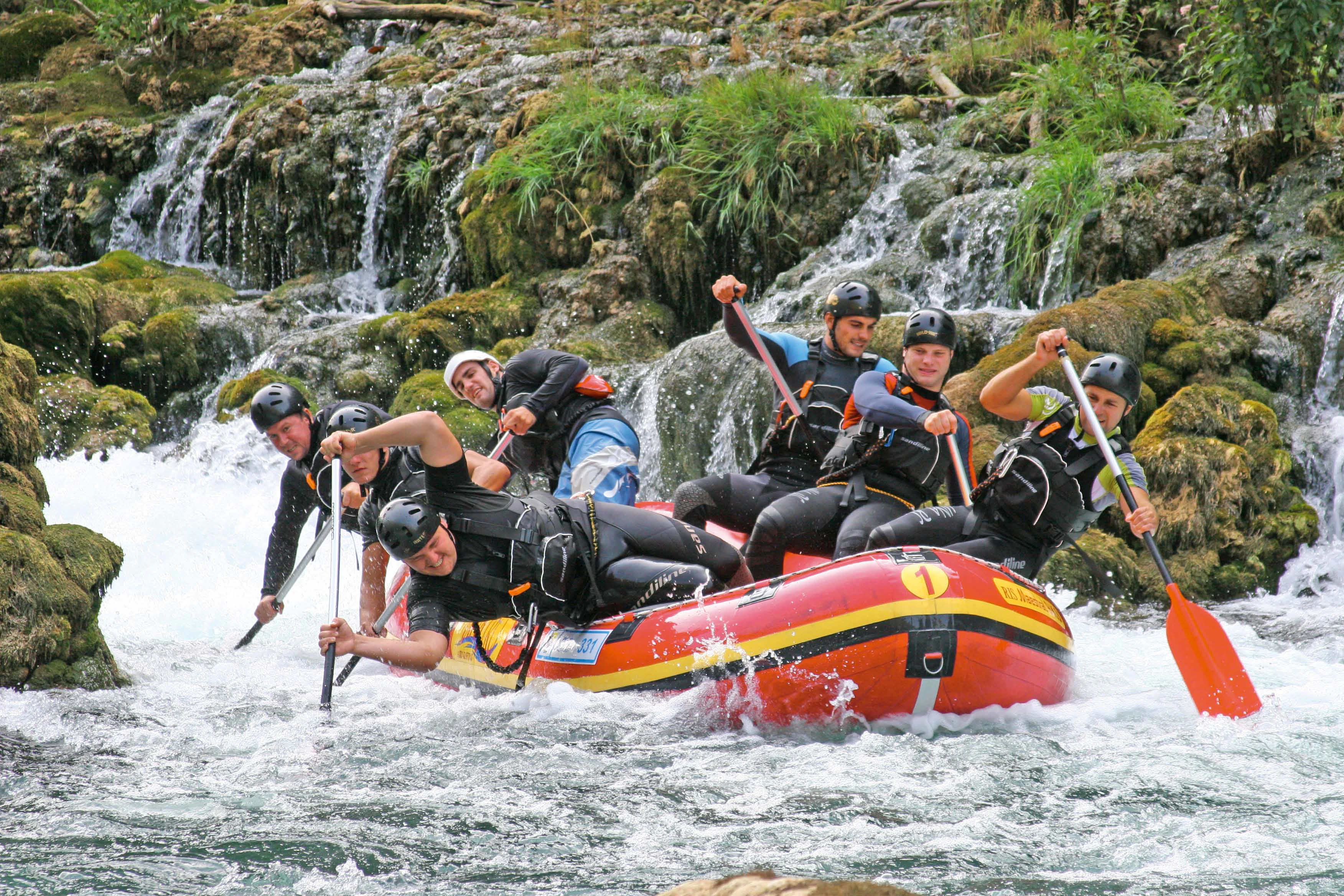 Group of people in a red inflatable raft navigating rapids on a river with waterfalls and rocky terrain