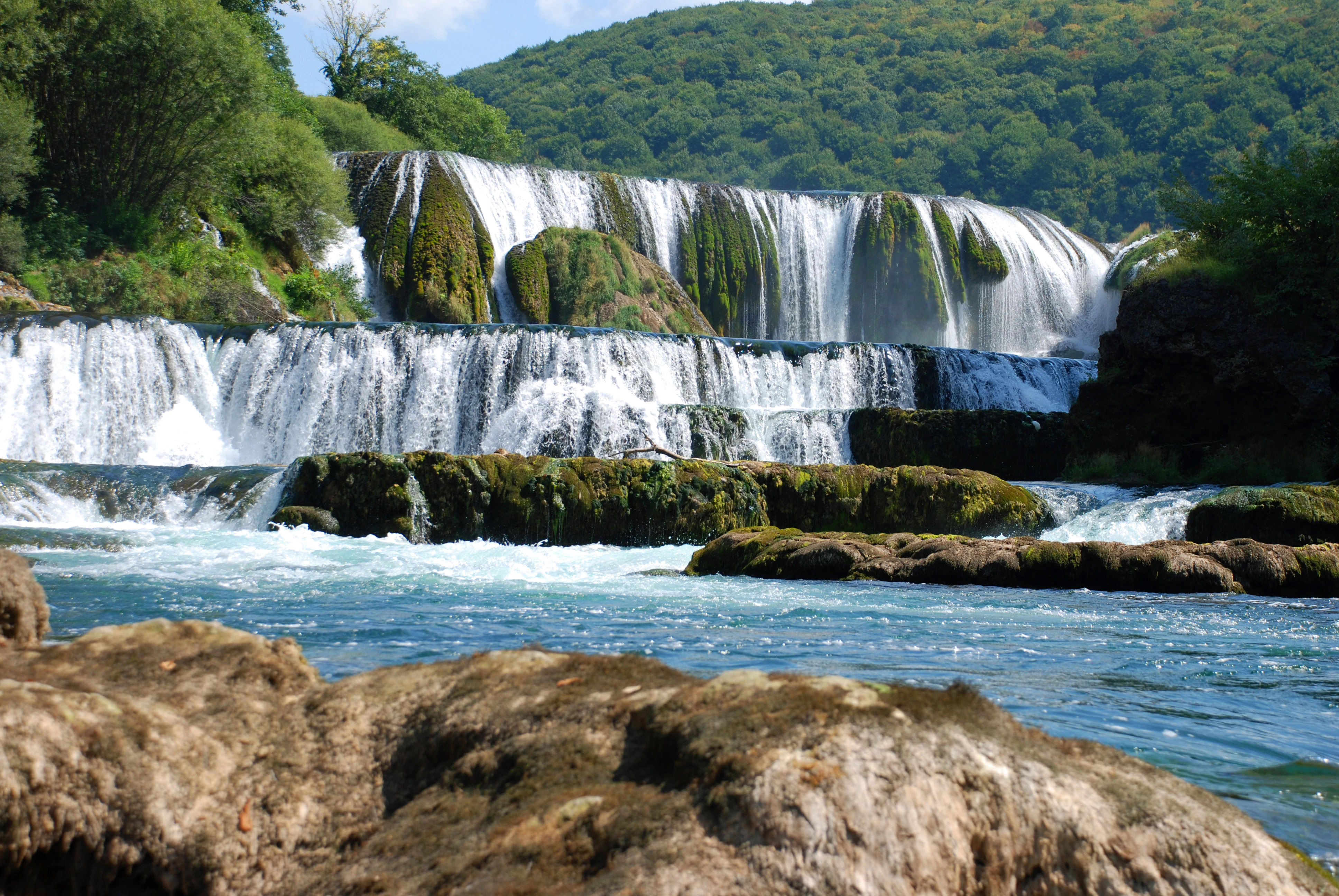 Multi-tiered waterfall flowing over moss-covered rocks with a blue river below and forested hills in the background