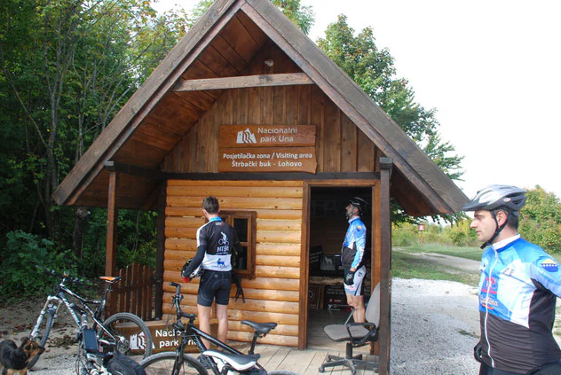 Wooden entrance structure with park signage, three cyclists in cycling gear, bicycles, and dogs in front of the building