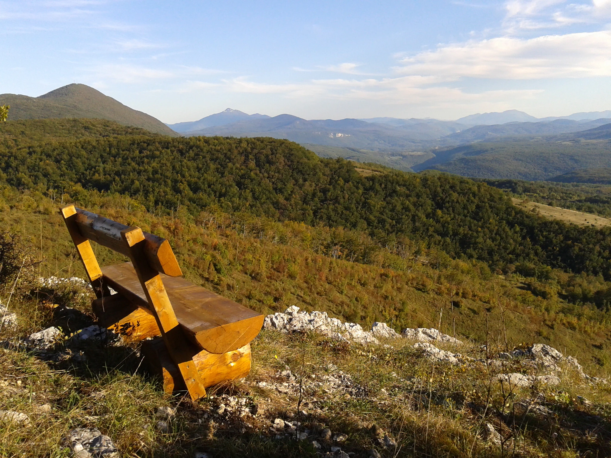 Wooden bench on rocky terrain overlooking a valley with forested mountains under a partly cloudy sky
