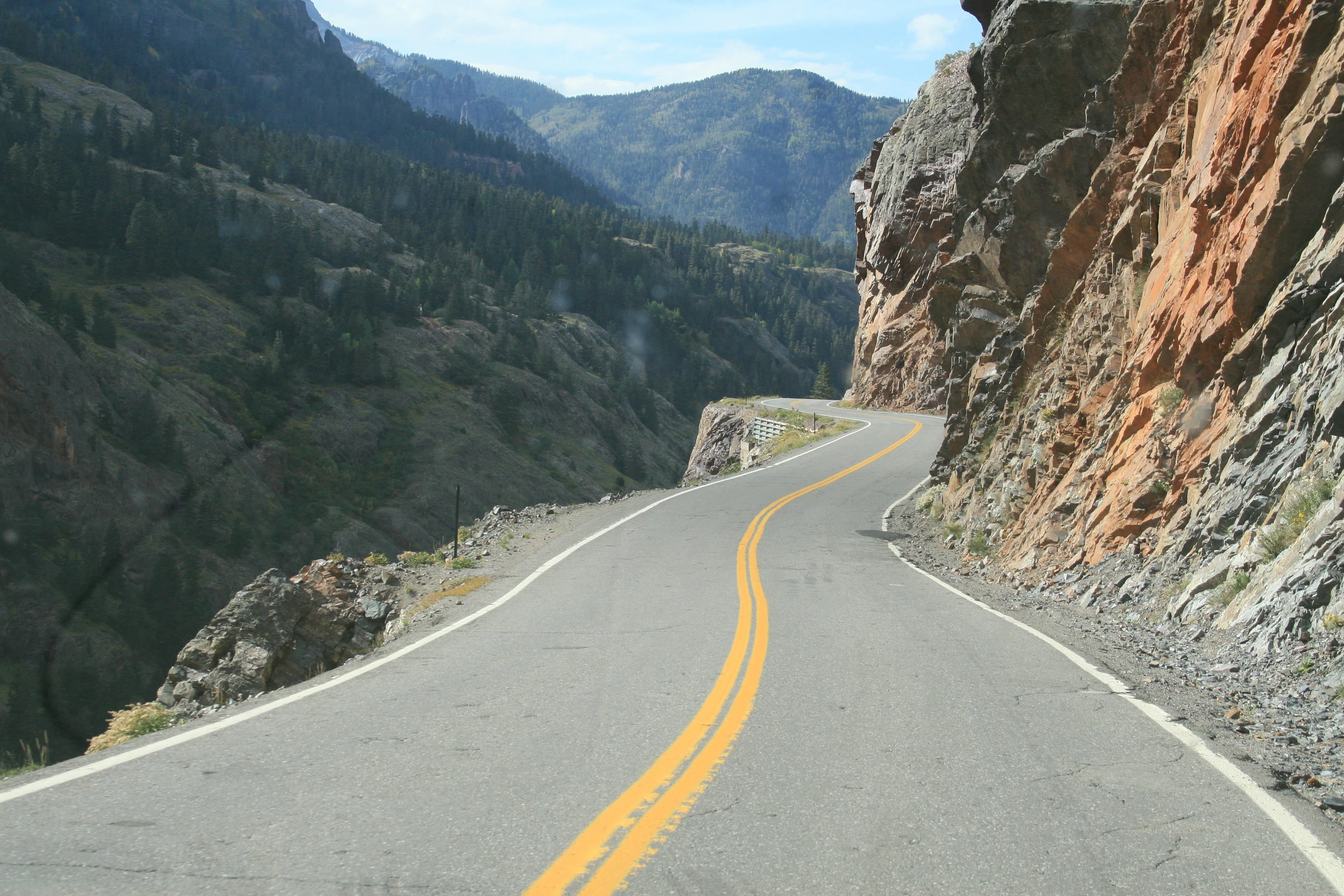A winding asphalt road with double yellow lines snakes through a steep rocky gorge with mountain slopes in the distance.
