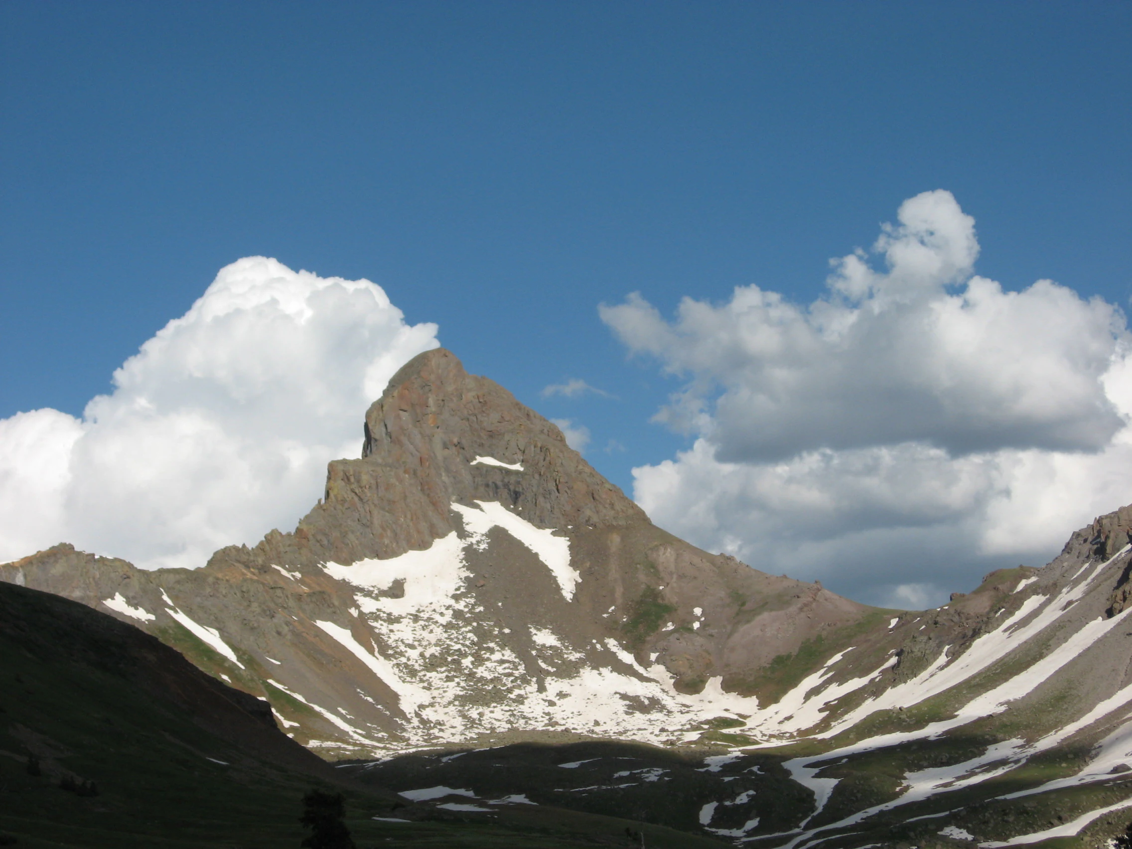 A jagged, triangular mountain peak with patches of snow on its steep slopes, set against a deep blue sky with white clouds.