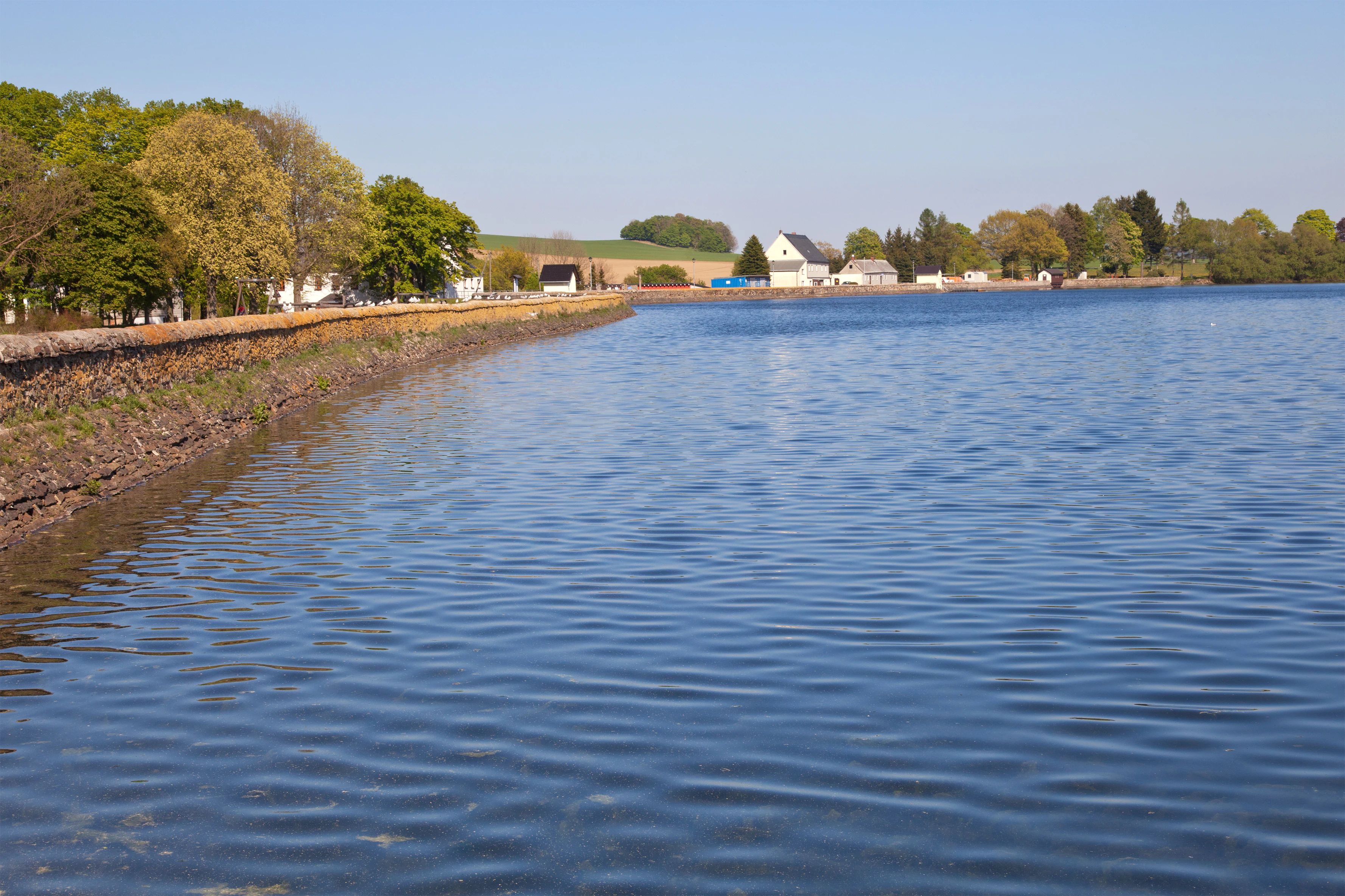 A wide angle view of a calm blue reservoir with a stone embankment wall on the left, leading to a small village with houses and trees in the distance under a clear sky.