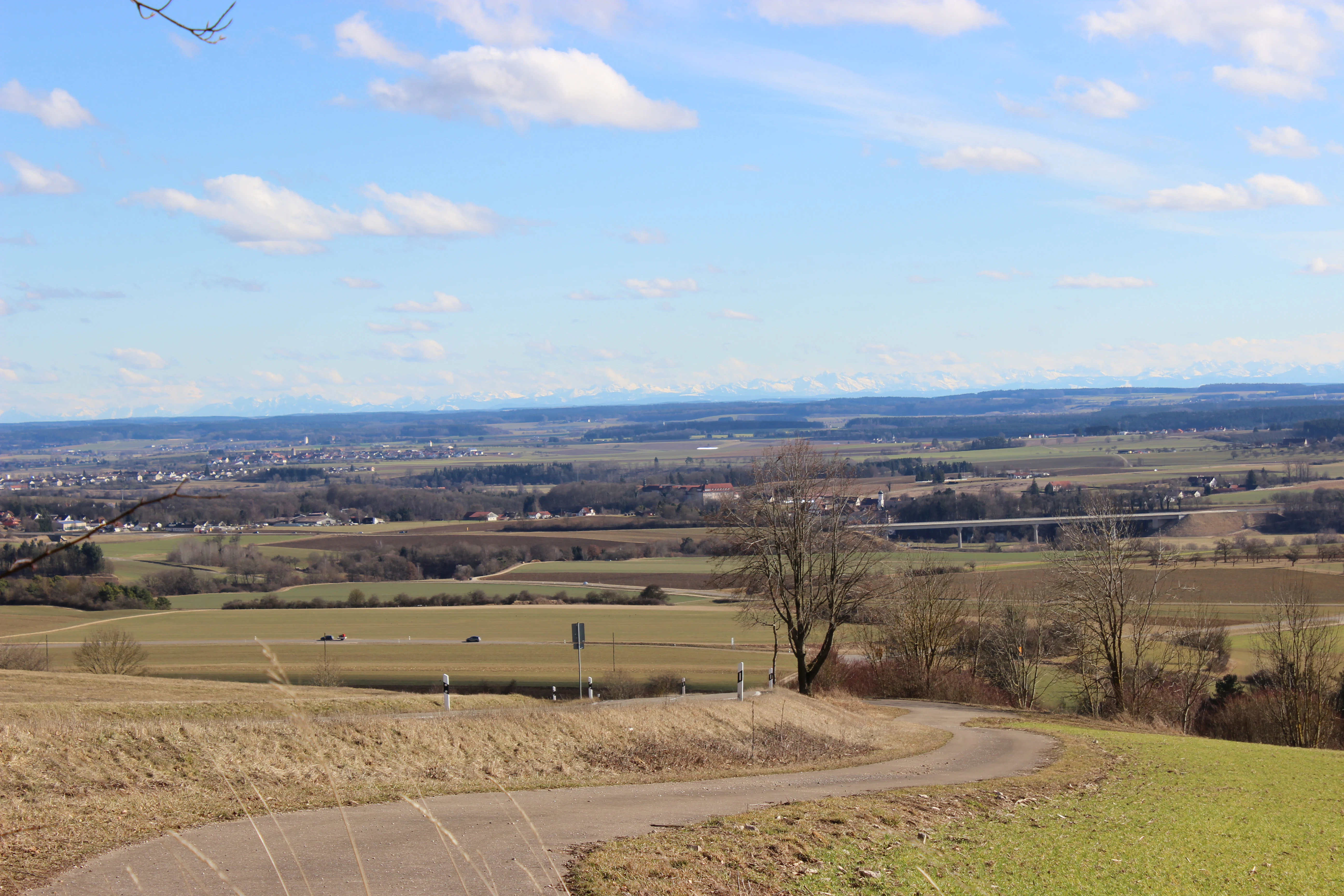 A wide landscape photograph from a high vantage point overlooking rolling fields and a small paved road in the foreground, with distant mountains visible on the horizon under a blue, partly cloudy sky.