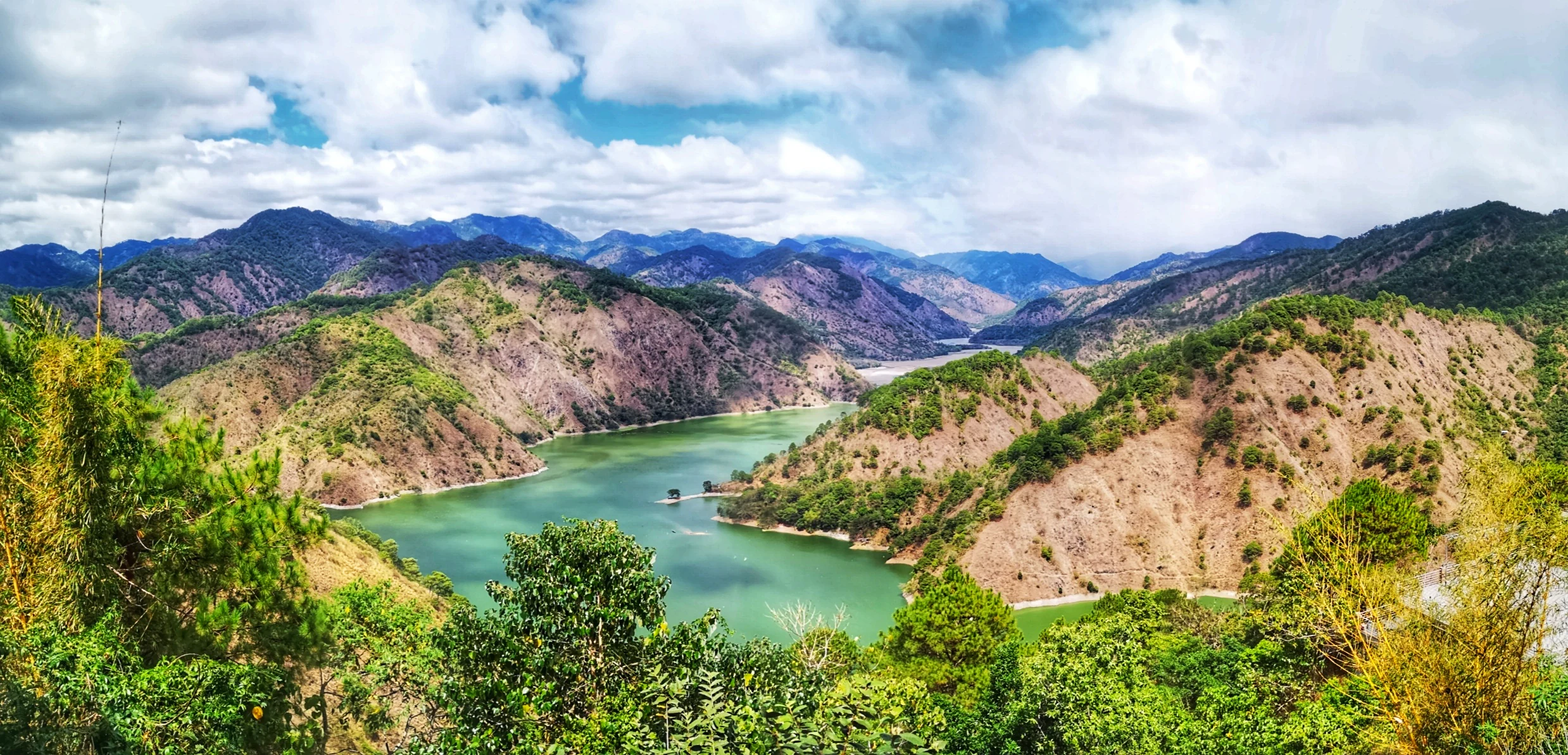 A wide panoramic landscape shot showing a winding green river reservoir surrounded by steep, rugged, and partially forested hills under a cloudy blue sky.